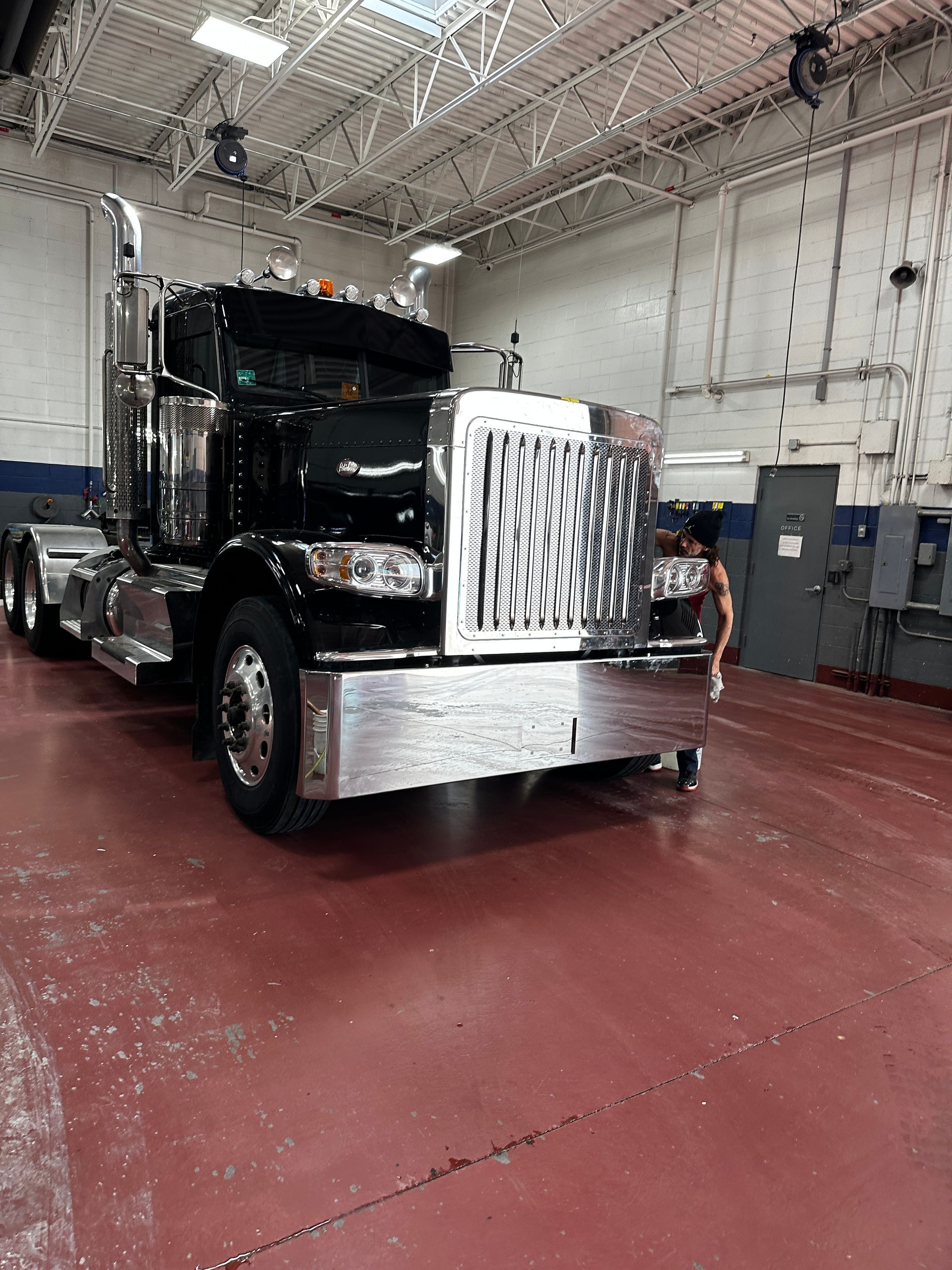 A black semi-truck with a polished chrome grille and bumper is parked inside a warehouse with a red floor.