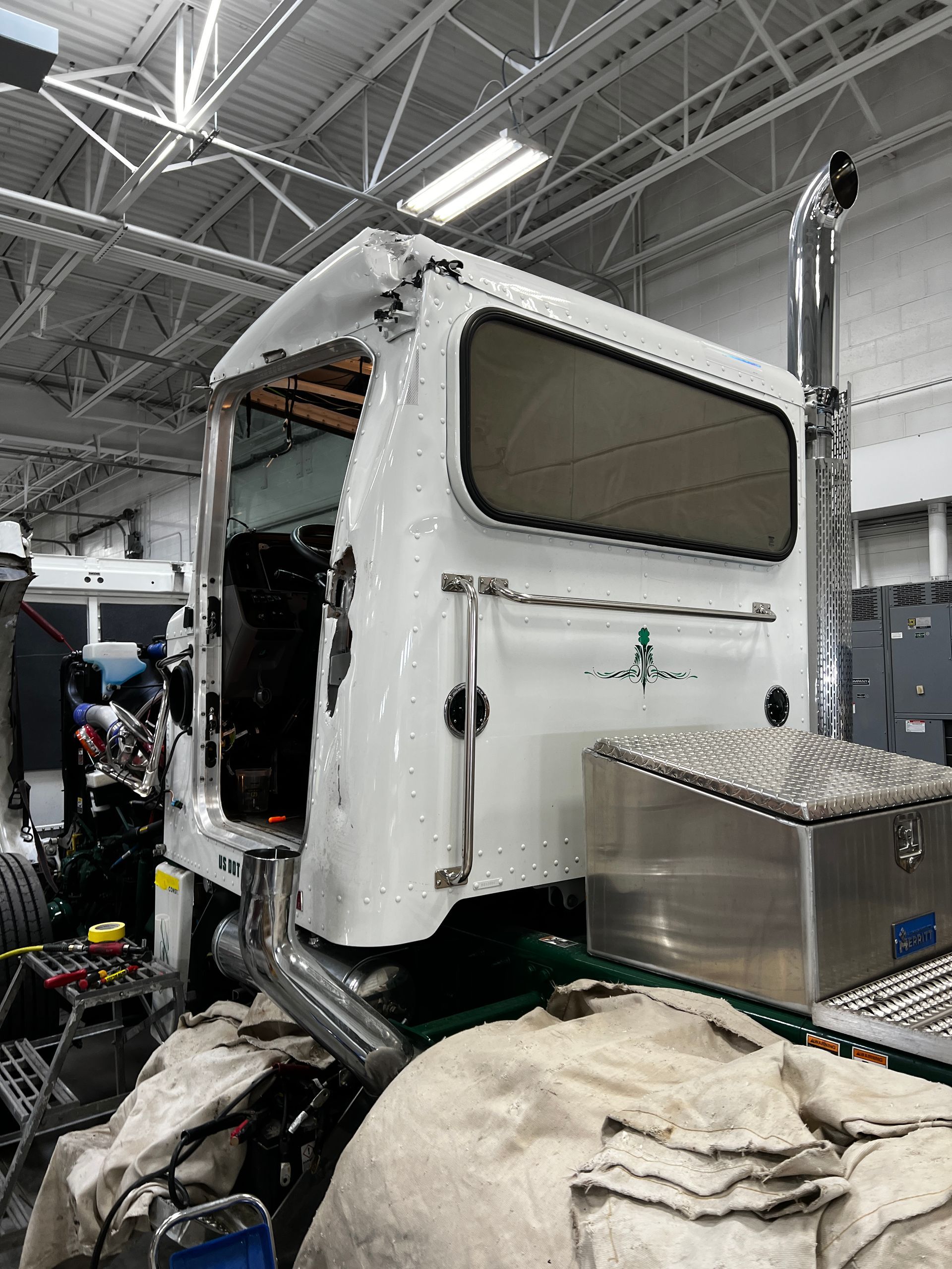 A white semi-truck cab in a workshop, featuring a chrome exhaust pipe, a side window, and a storage box on the chassis.