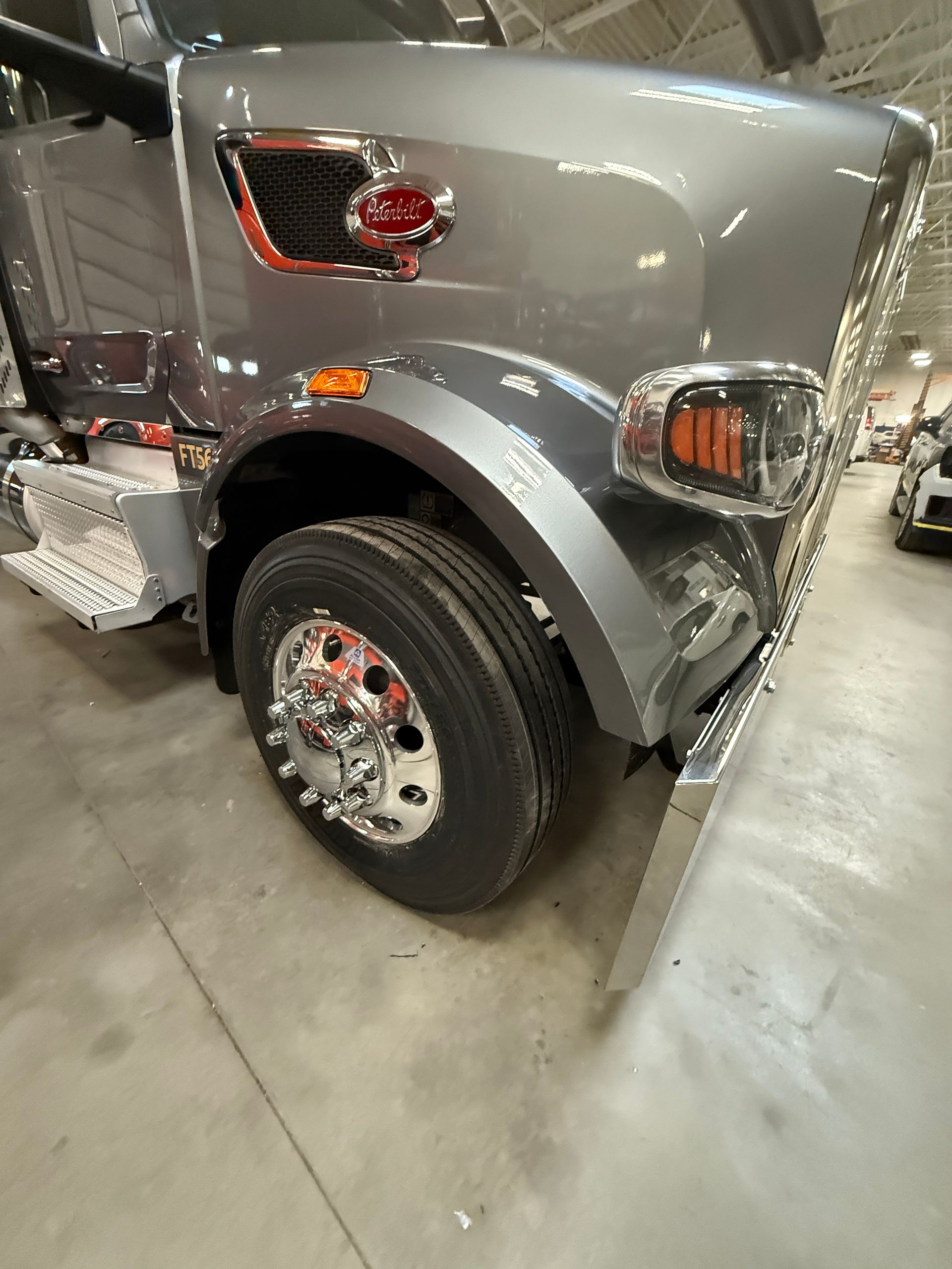 A close-up, angled view of the front passenger side of a silver semi-truck parked on a concrete warehouse floor.