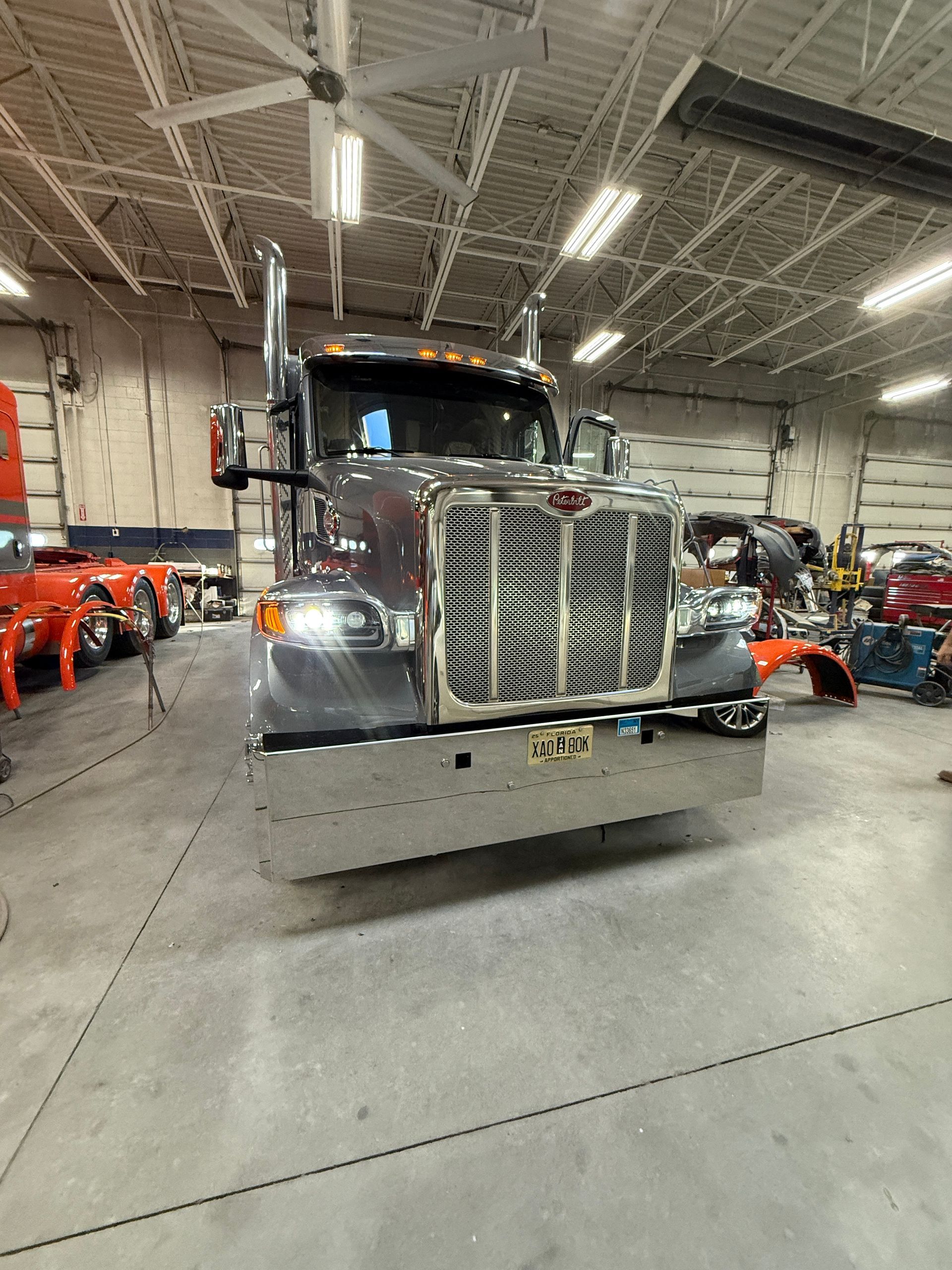 A grey semi-truck with a shiny chrome bumper and large mesh grille parked inside a well-lit repair shop.