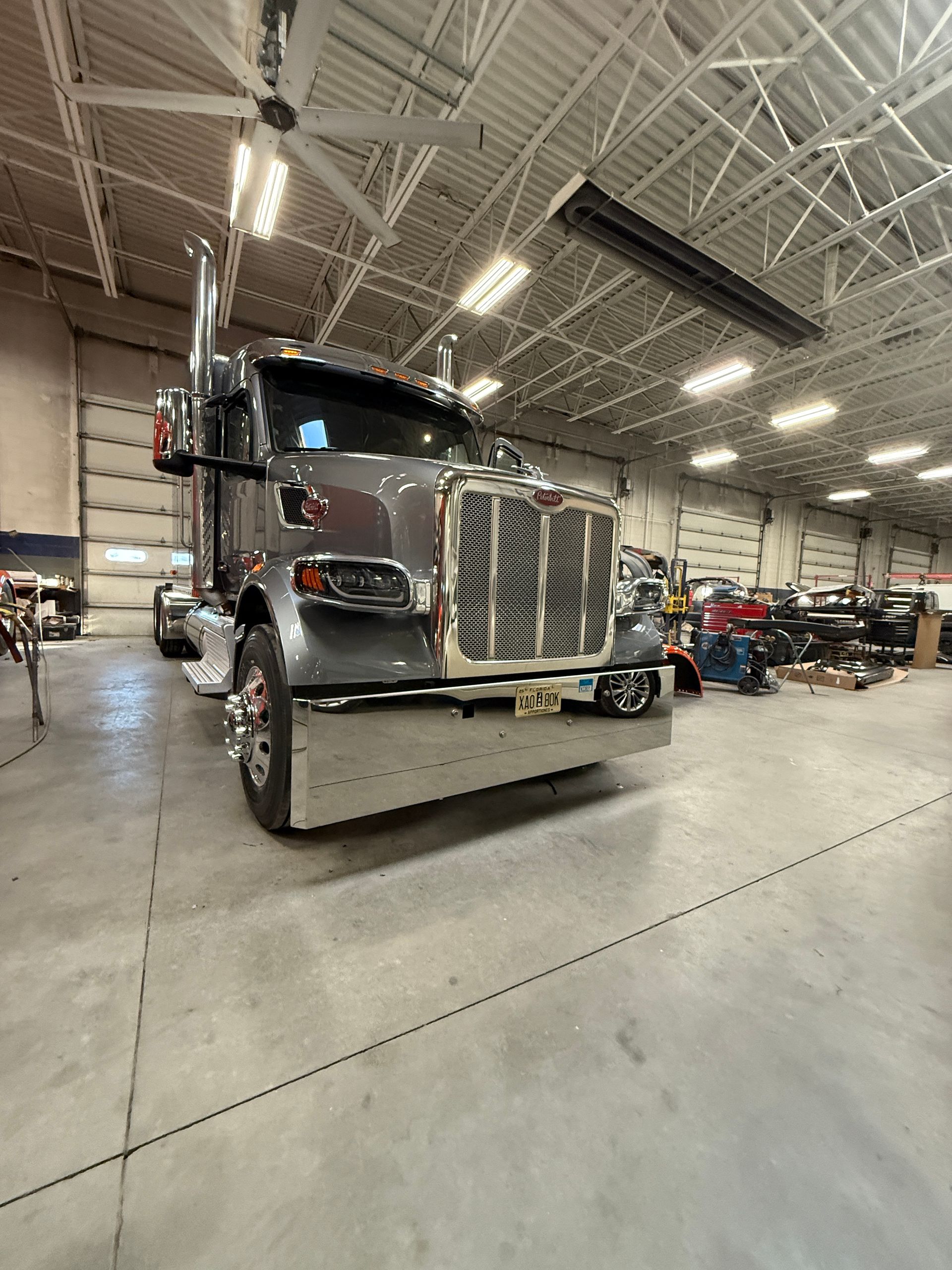 A gray Peterbilt semi-truck with a large chrome bumper parked inside a brightly lit, spacious maintenance garage.
