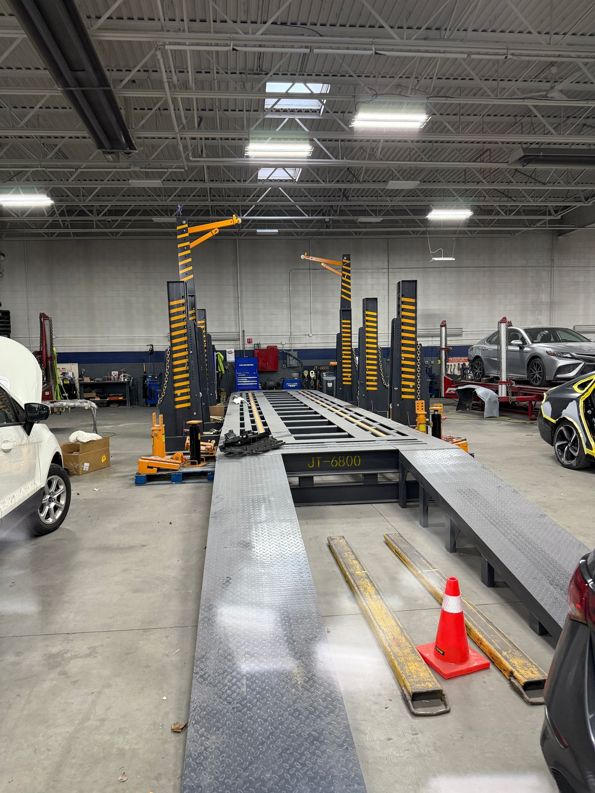 A grey automotive repair shop floor featuring a large black frame rack for vehicle alignment and a lone orange safety cone.