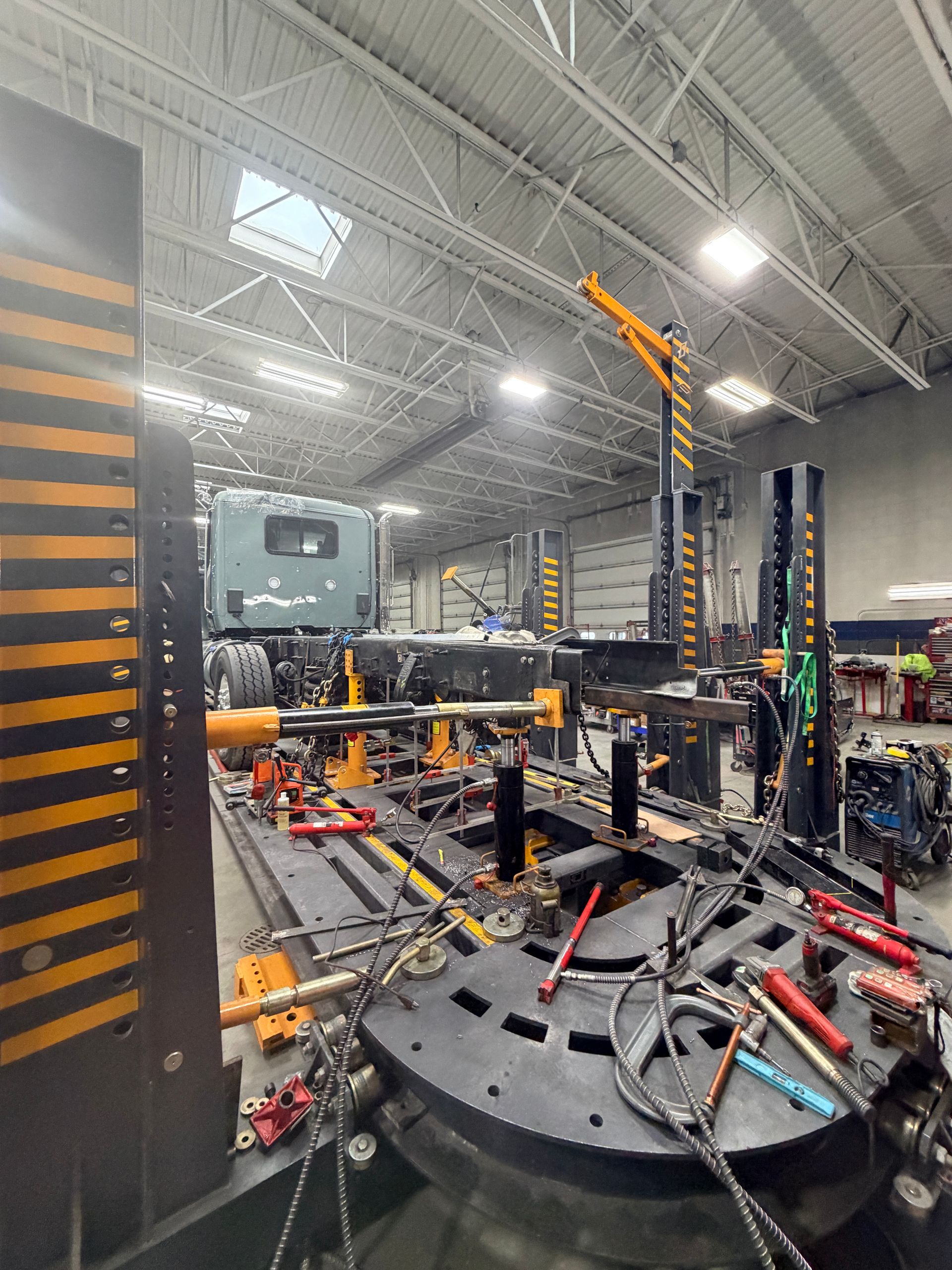 A truck chassis sits on an industrial frame machine inside a workshop for repair, surrounded by metal tools and equipment.