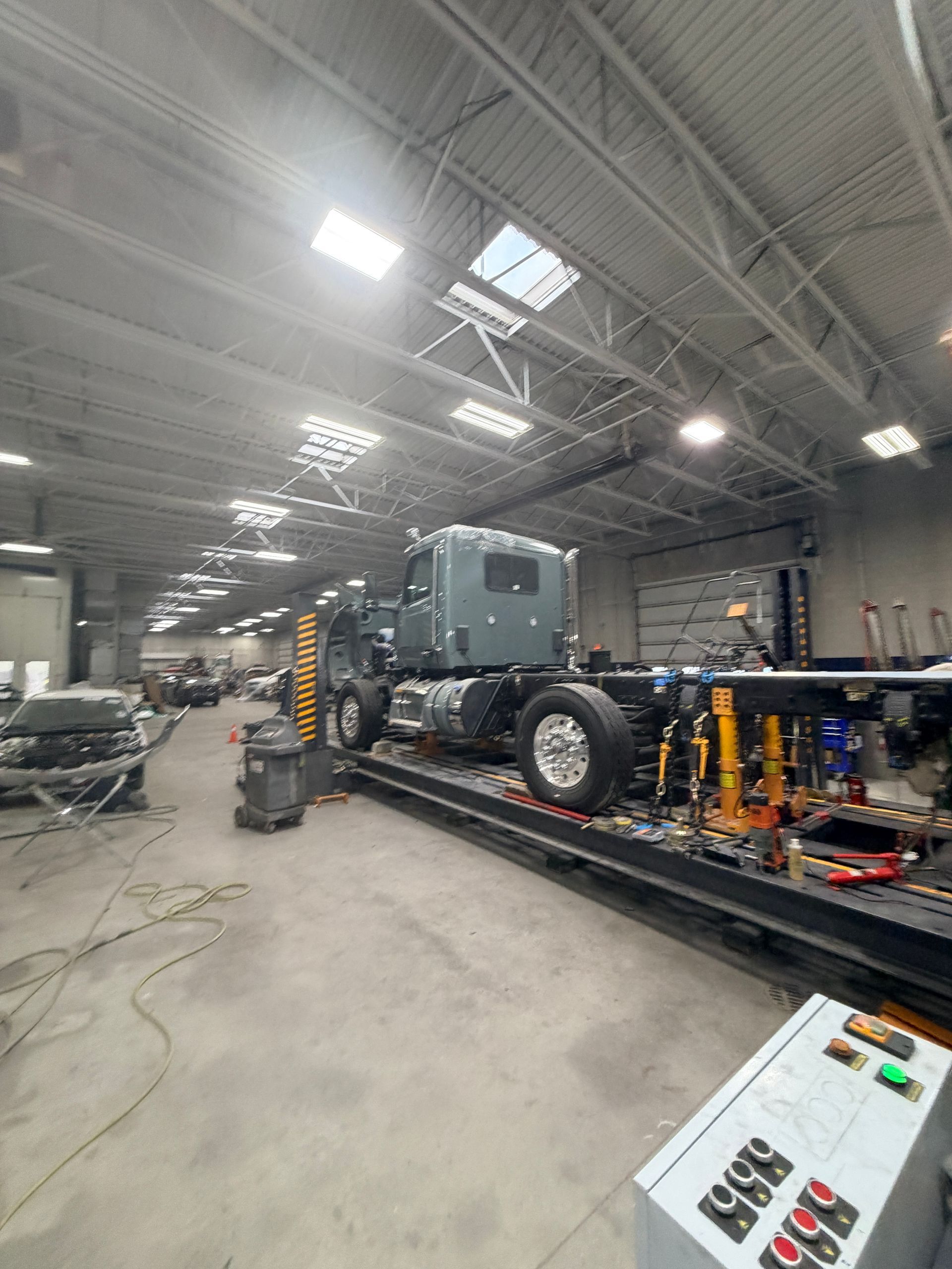 A gray semi-truck cab sits on an industrial frame rack inside a large, brightly lit automotive workshop.
