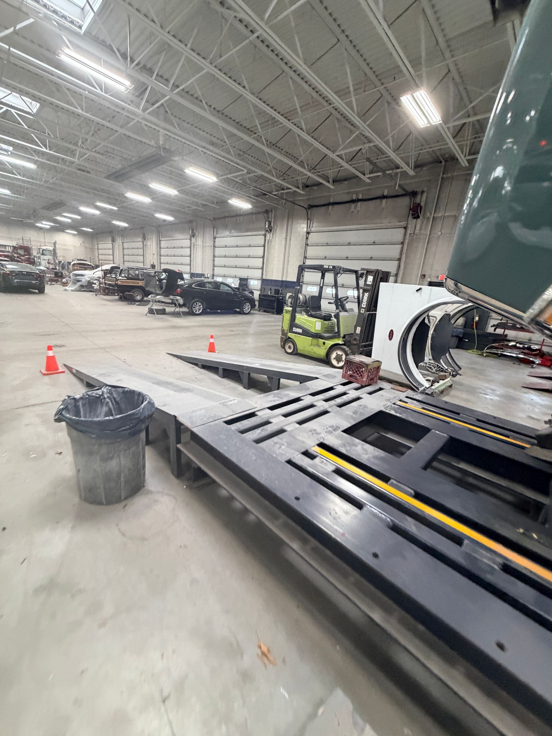 A warehouse showing a black car hauler ramp, a green forklift, and parked vehicles under bright ceiling lights.