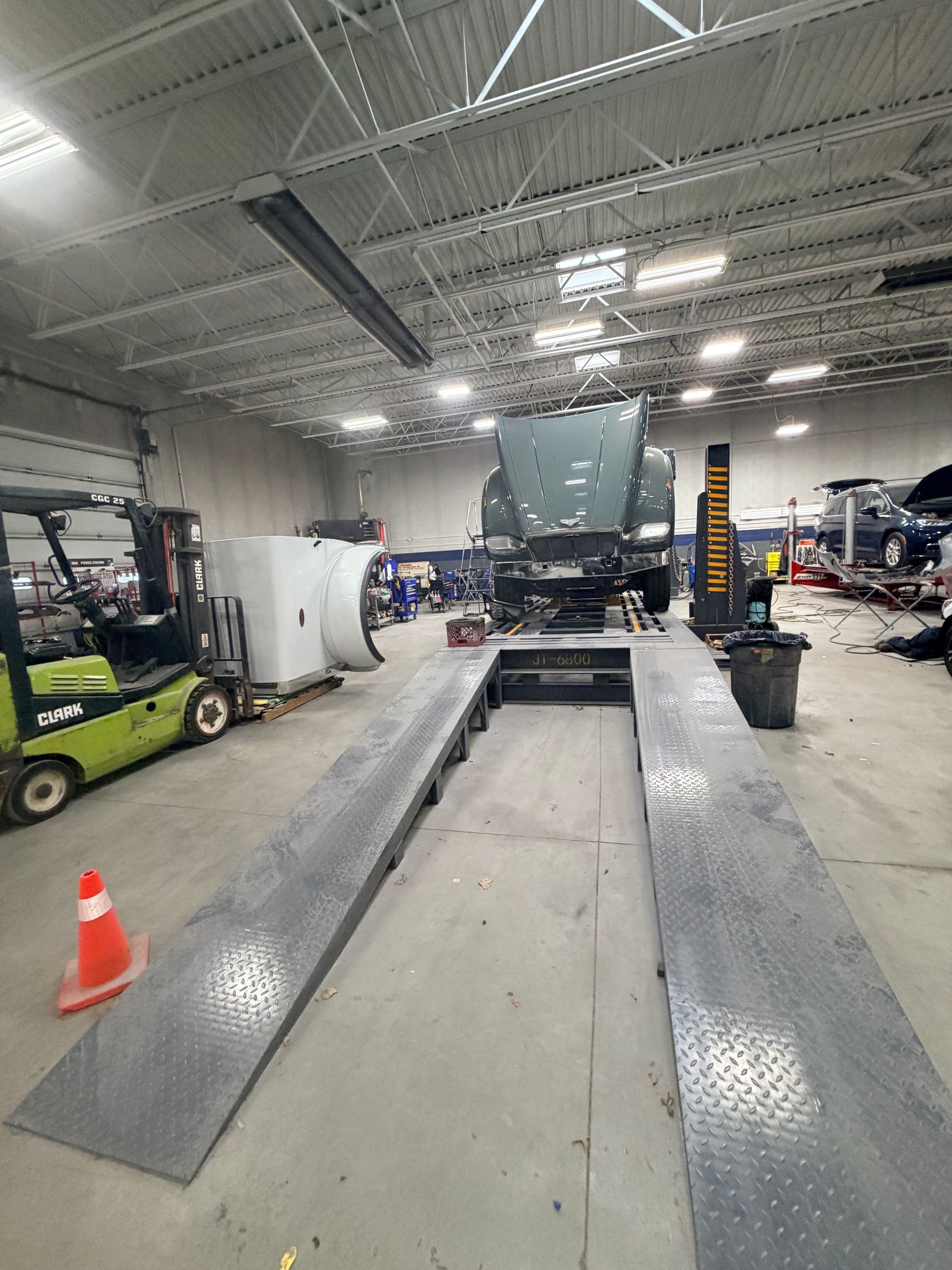 A semi-truck parked on a metal inspection ramp inside a brightly lit industrial repair shop with a forklift to the left.
