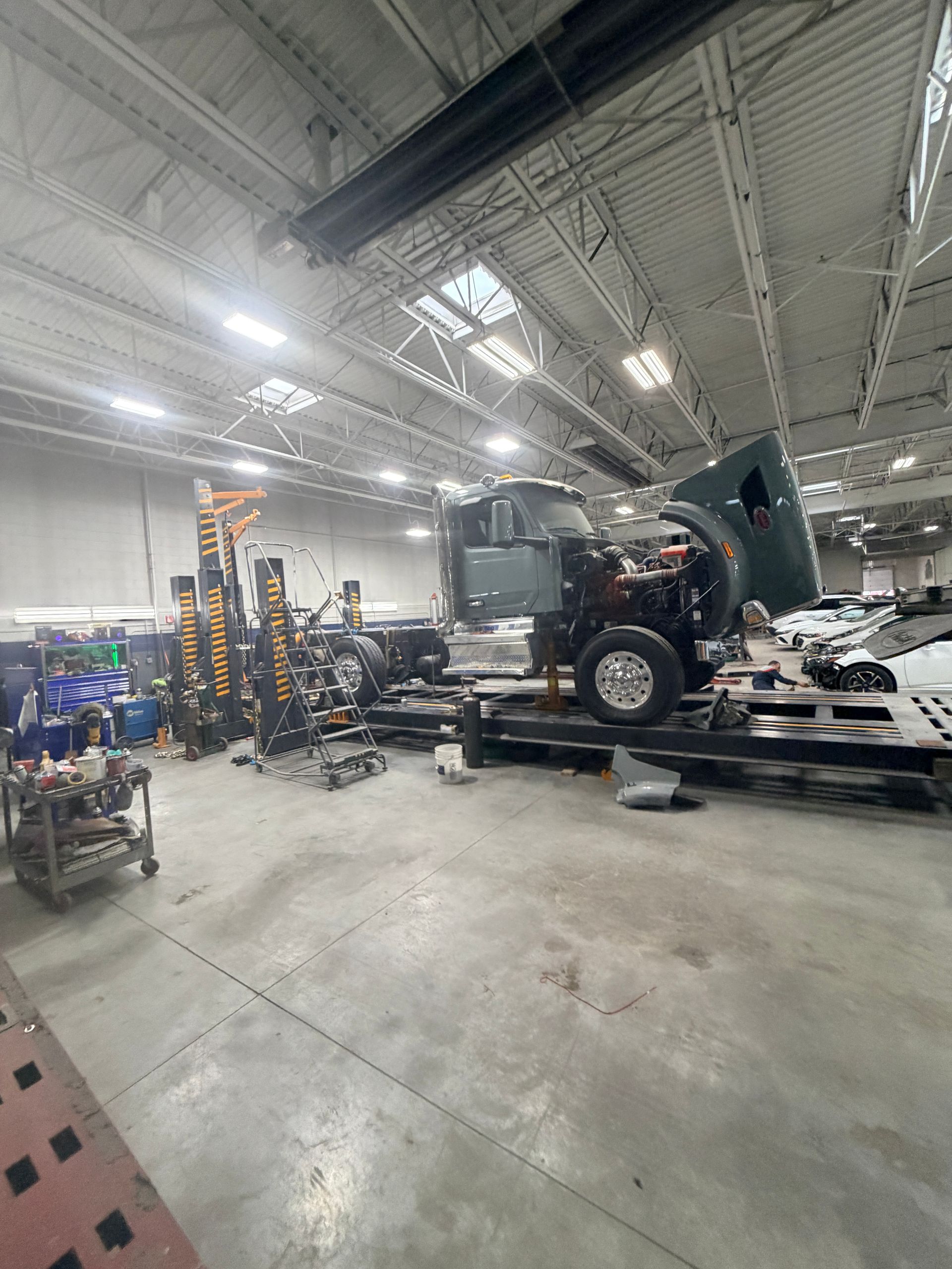 A large semi-truck with its hood raised sits on a service lift inside an industrial repair garage.