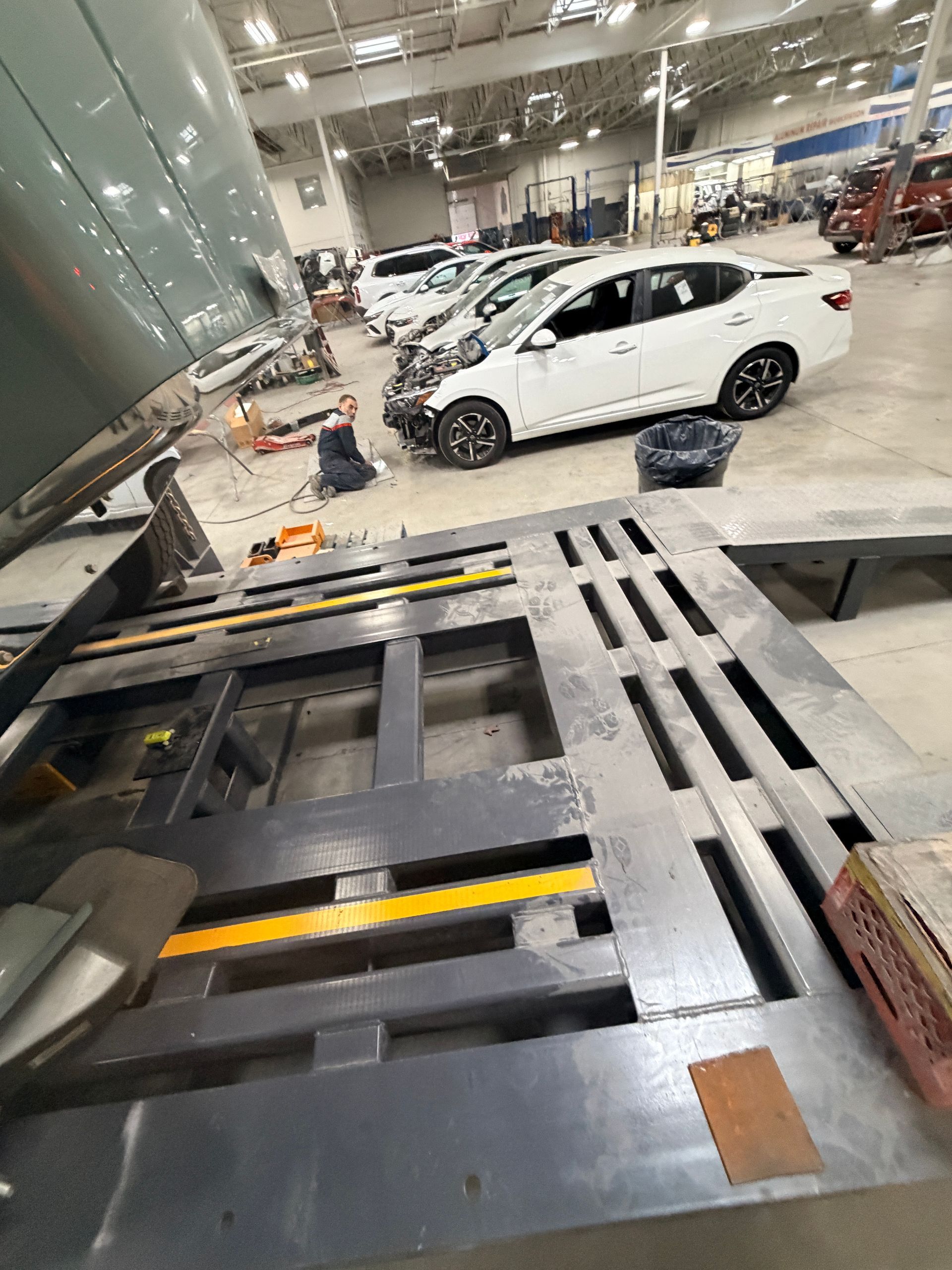 Inside an auto repair shop, a mechanic works on a white sedan on a frame rack near a raised, heavy-duty vehicle lift.
