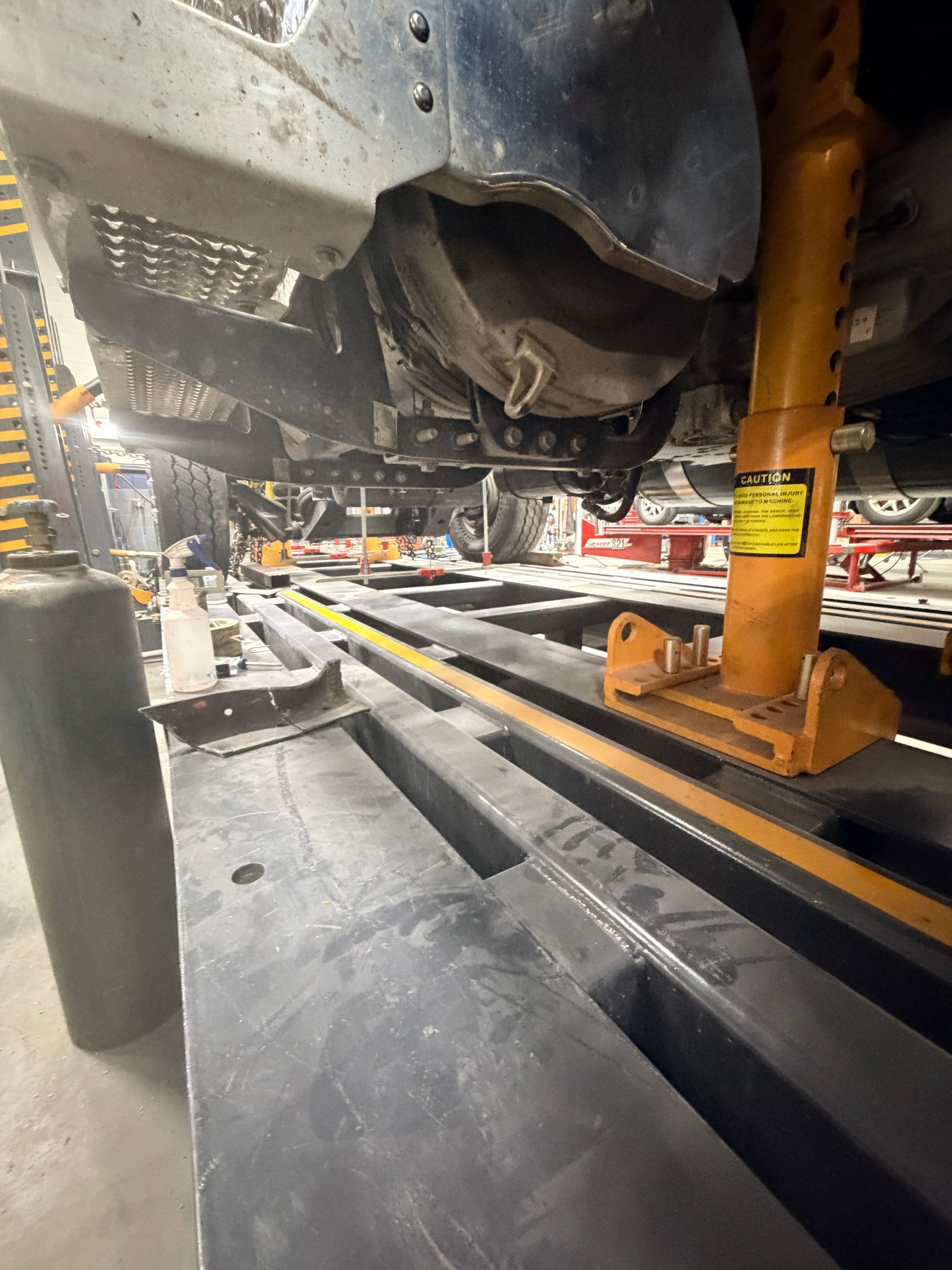 A vehicle is raised on a metal lift in an auto repair shop, with an orange jack stand supporting the frame.