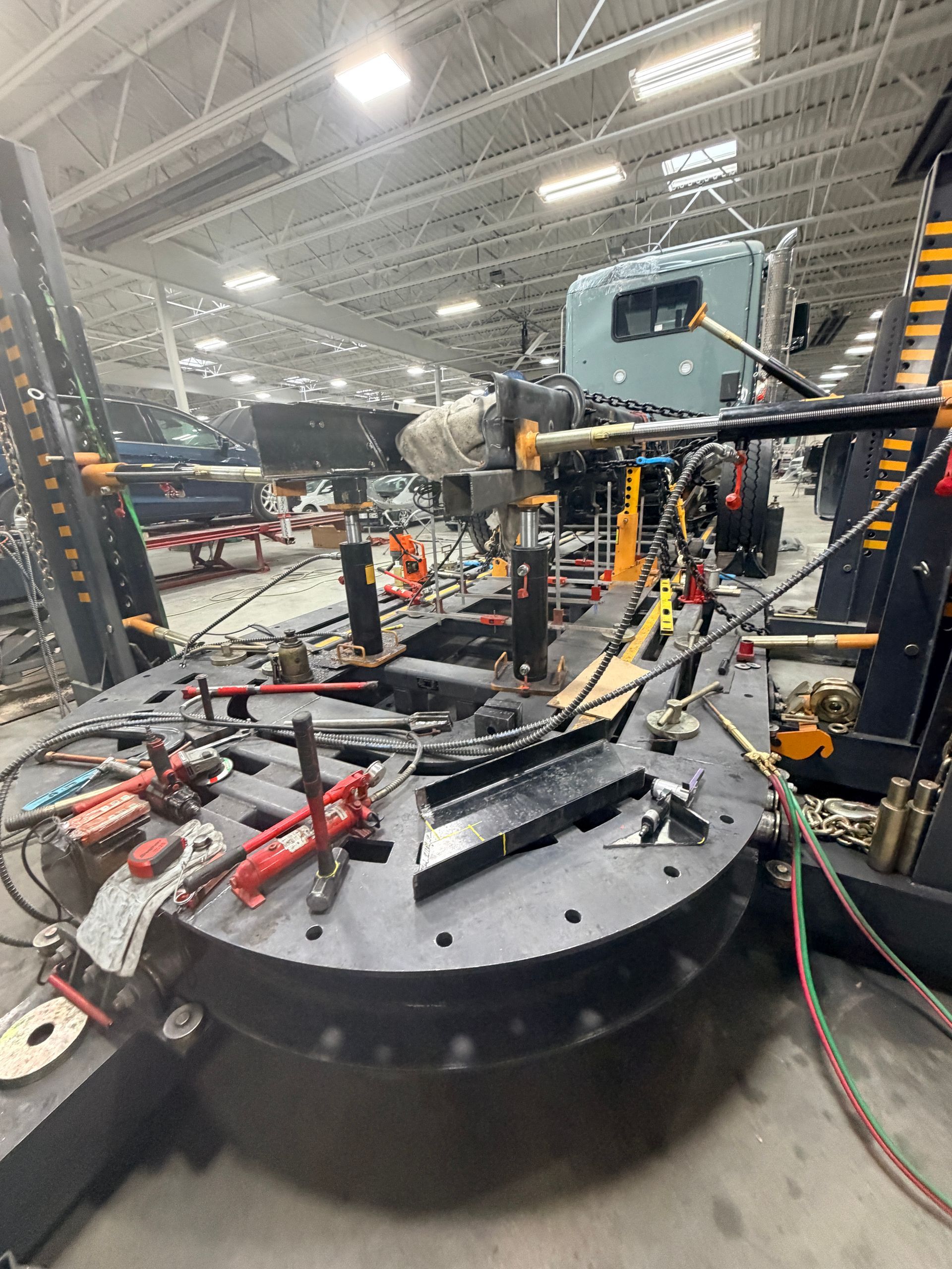 A truck frame sits on a heavy-duty industrial frame straightener rack inside a large, bright repair shop.