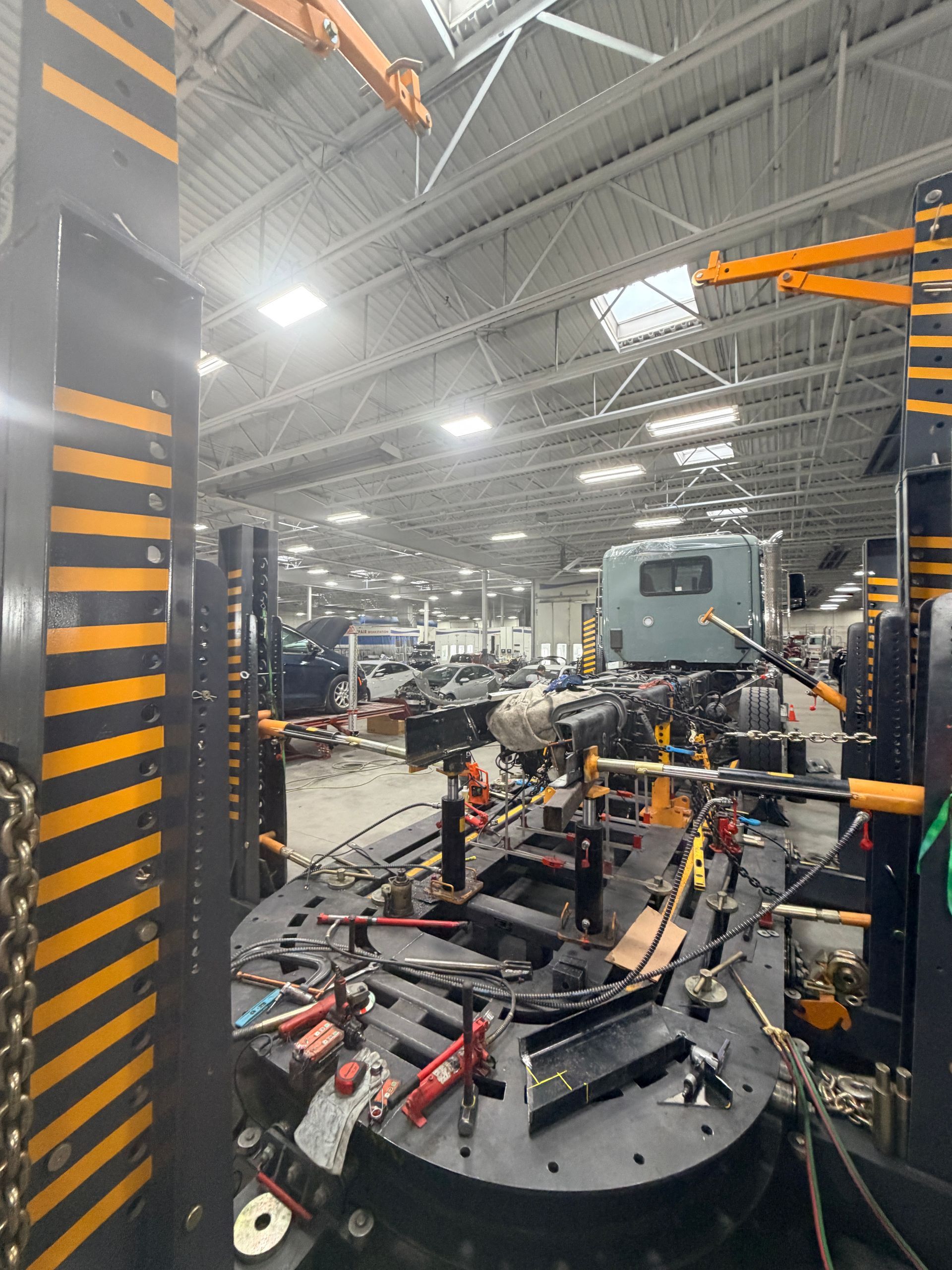 A gray truck cab positioned on an industrial frame repair rack inside a large, brightly lit workshop.