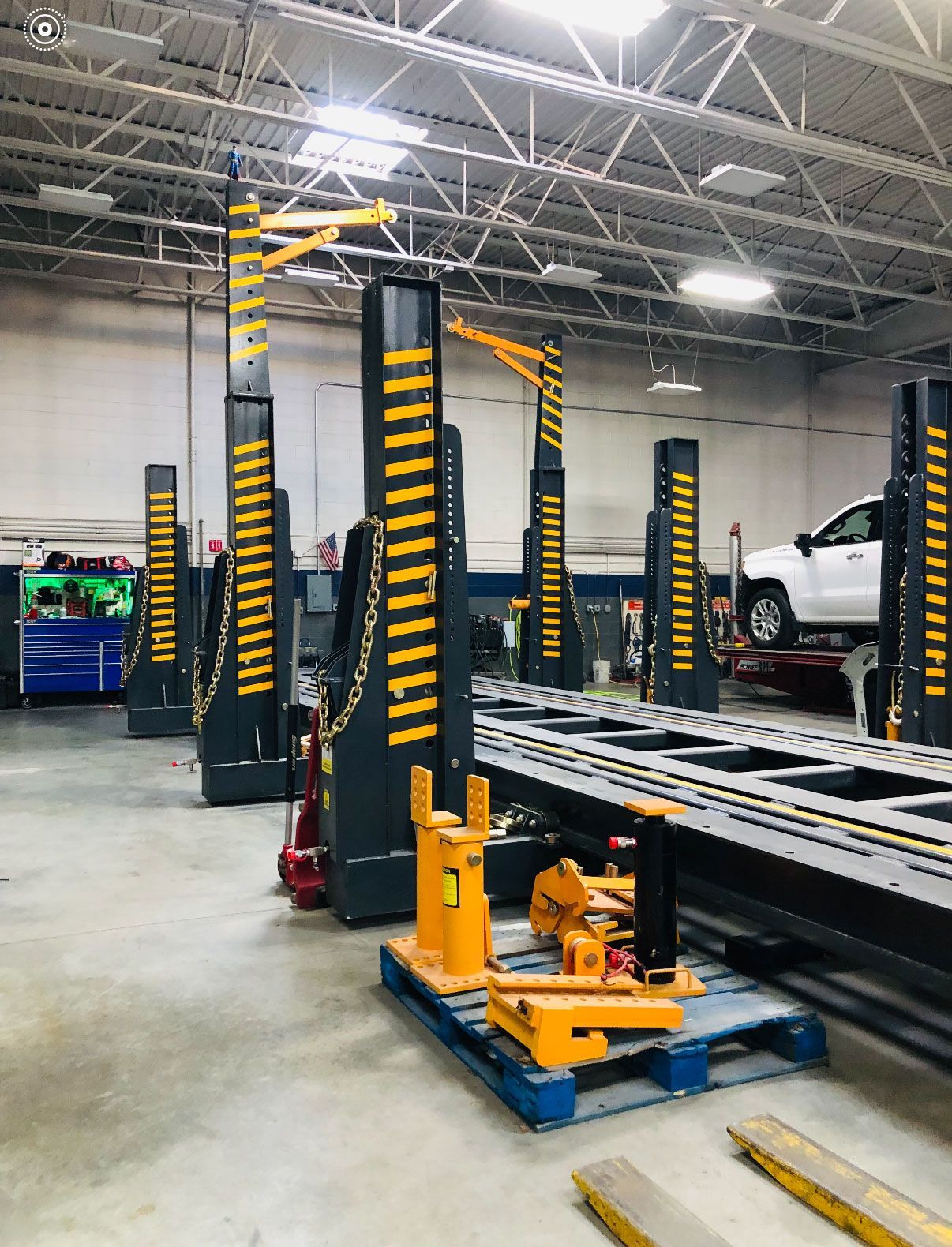 Heavy-duty vehicle lift columns in a workshop, with components on a pallet and a car on a lift in the background.