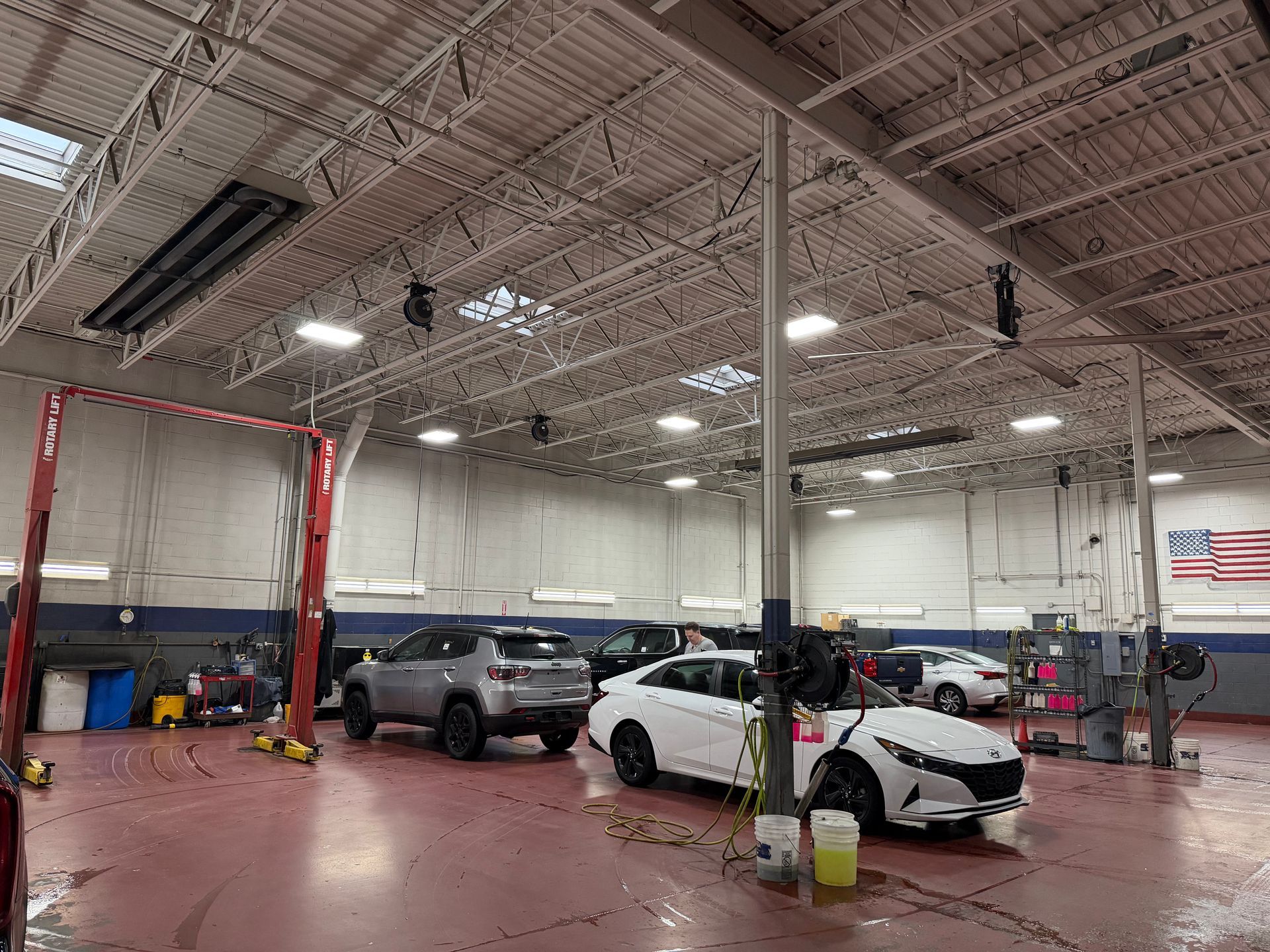 Automotive repair shop featuring a gray SUV and white sedan parked on a red floor beneath an exposed ceiling structure.