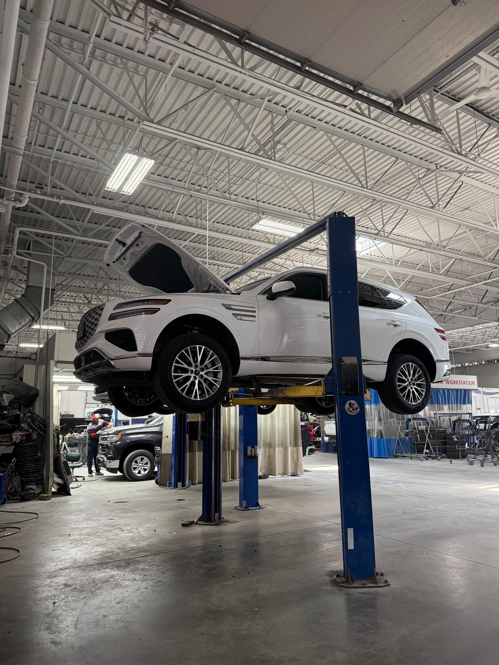 A white SUV with its hood raised is lifted on a two-post hydraulic lift inside a brightly lit automotive repair shop.