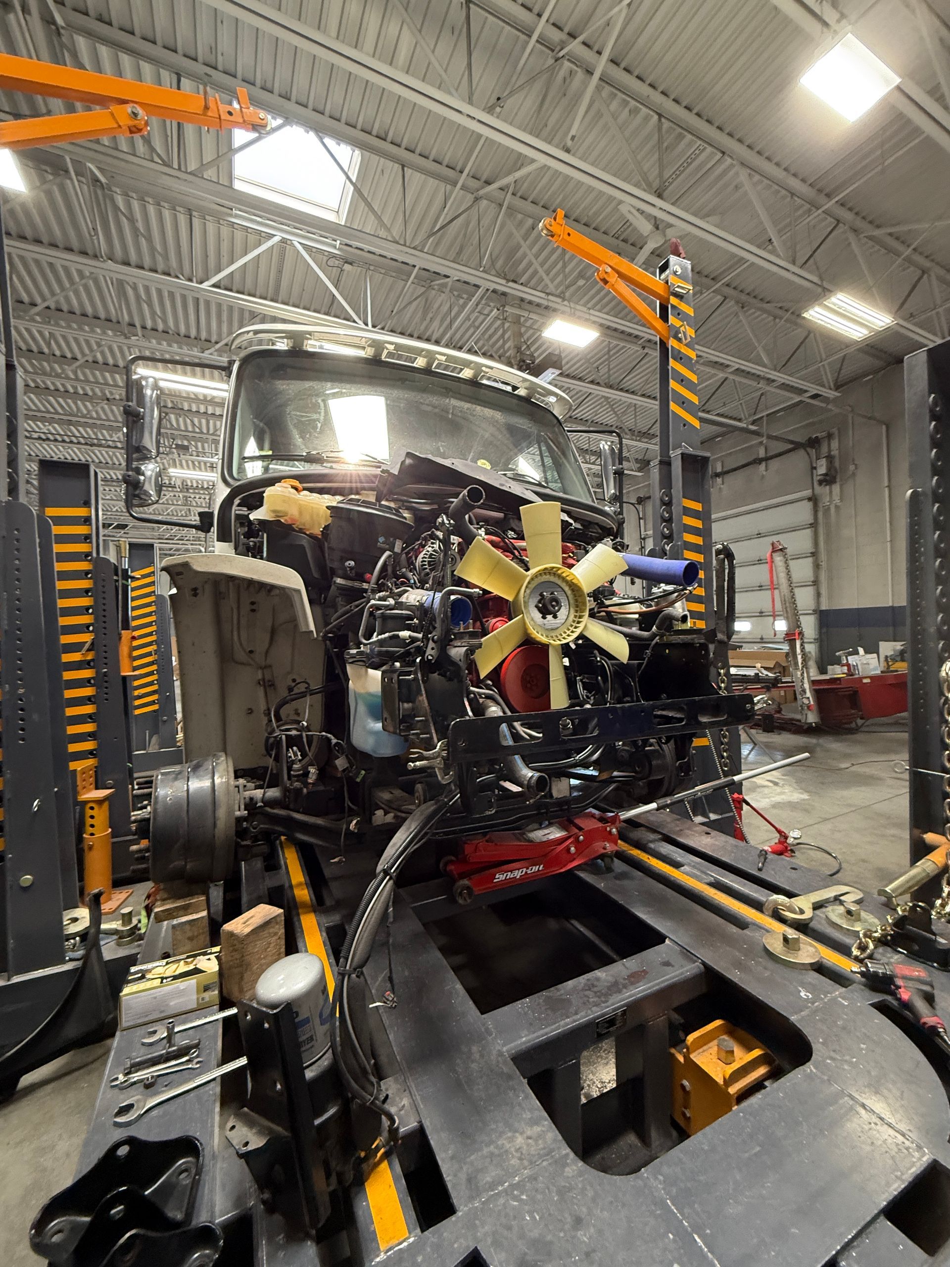 A truck engine with a large yellow cooling fan being serviced in an industrial workshop with vehicle lifts.