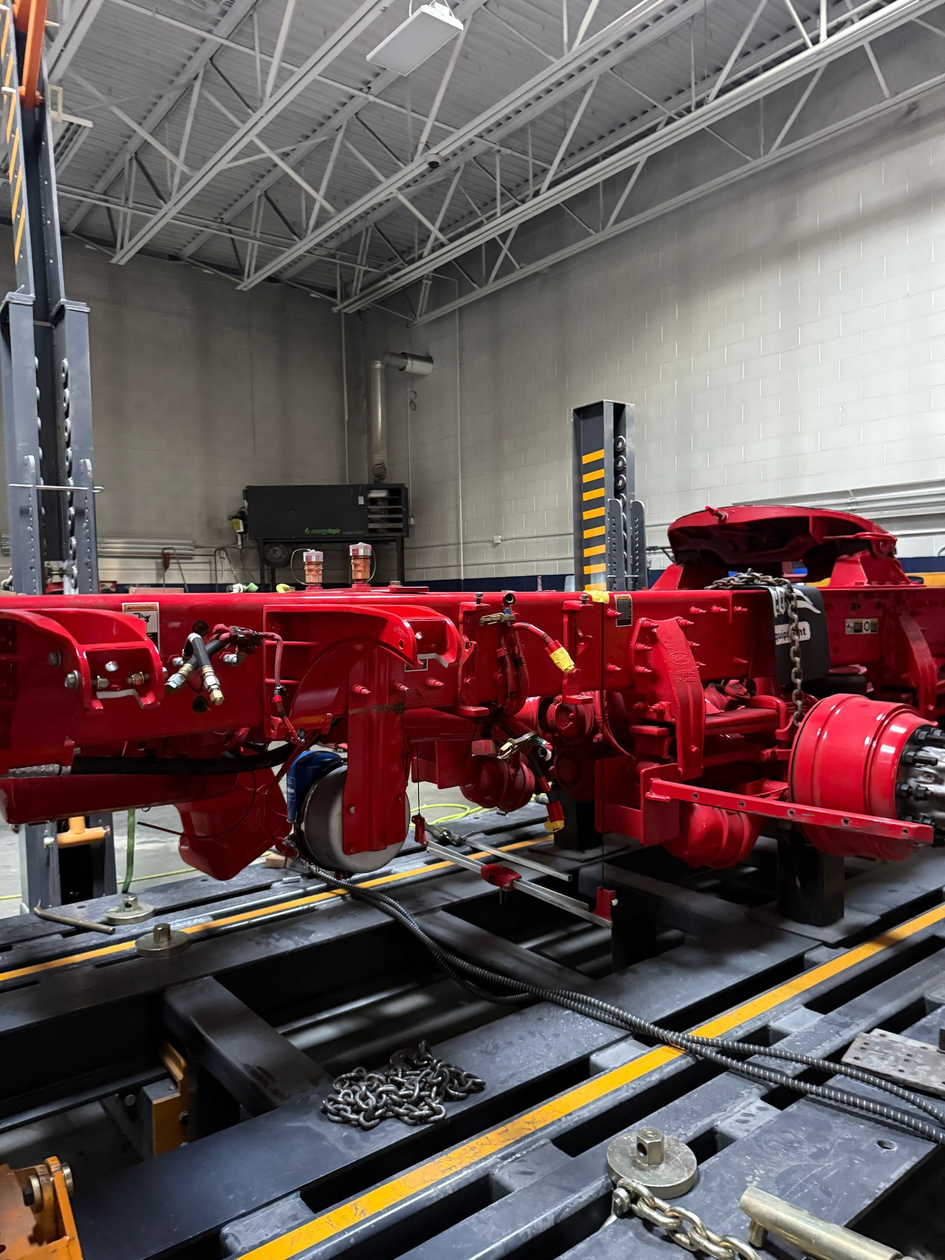 A red truck chassis sits on an elevated platform in a workshop, suspended by heavy-duty lifting equipment.