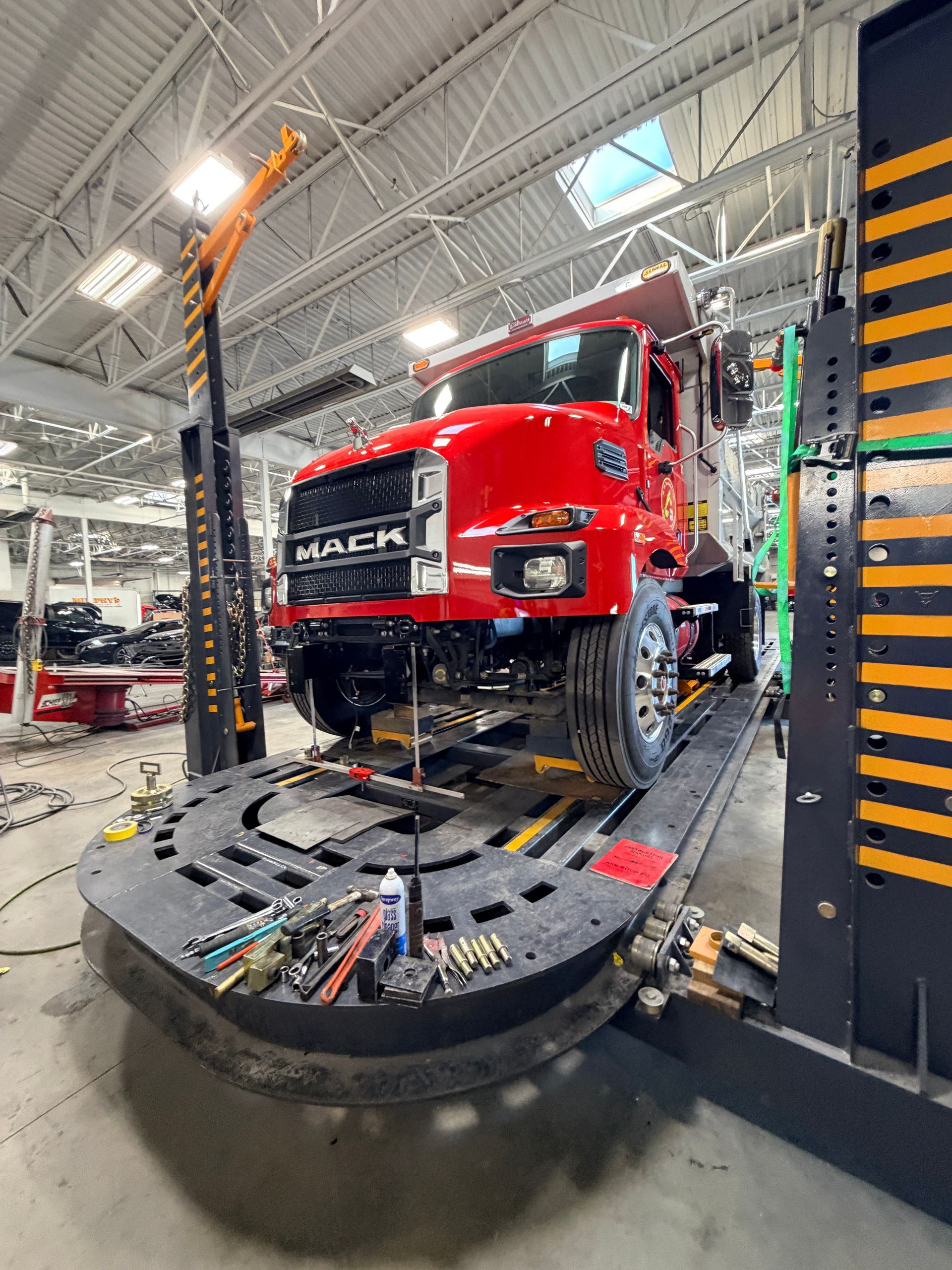 A bright red Mack dump truck is raised on a heavy-duty alignment rack in an industrial workshop.