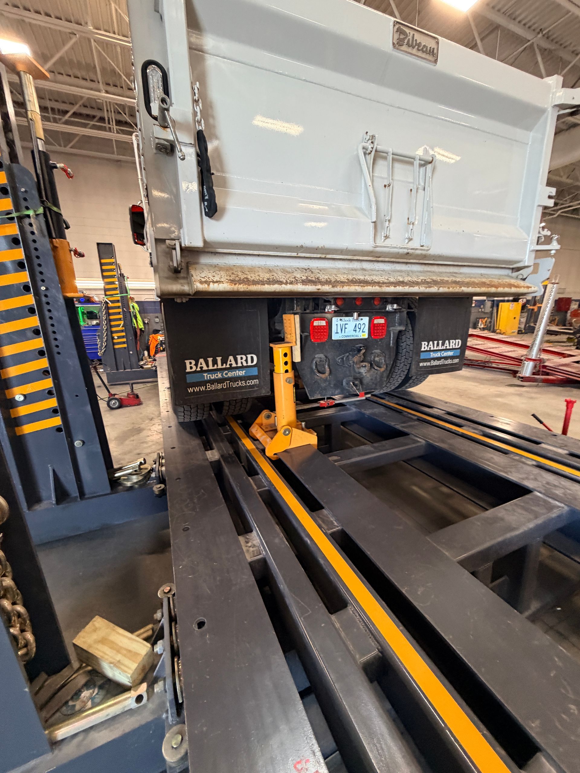 A white dump truck is positioned over a heavy-duty industrial service lift inside a garage, supported by yellow jack stands.
