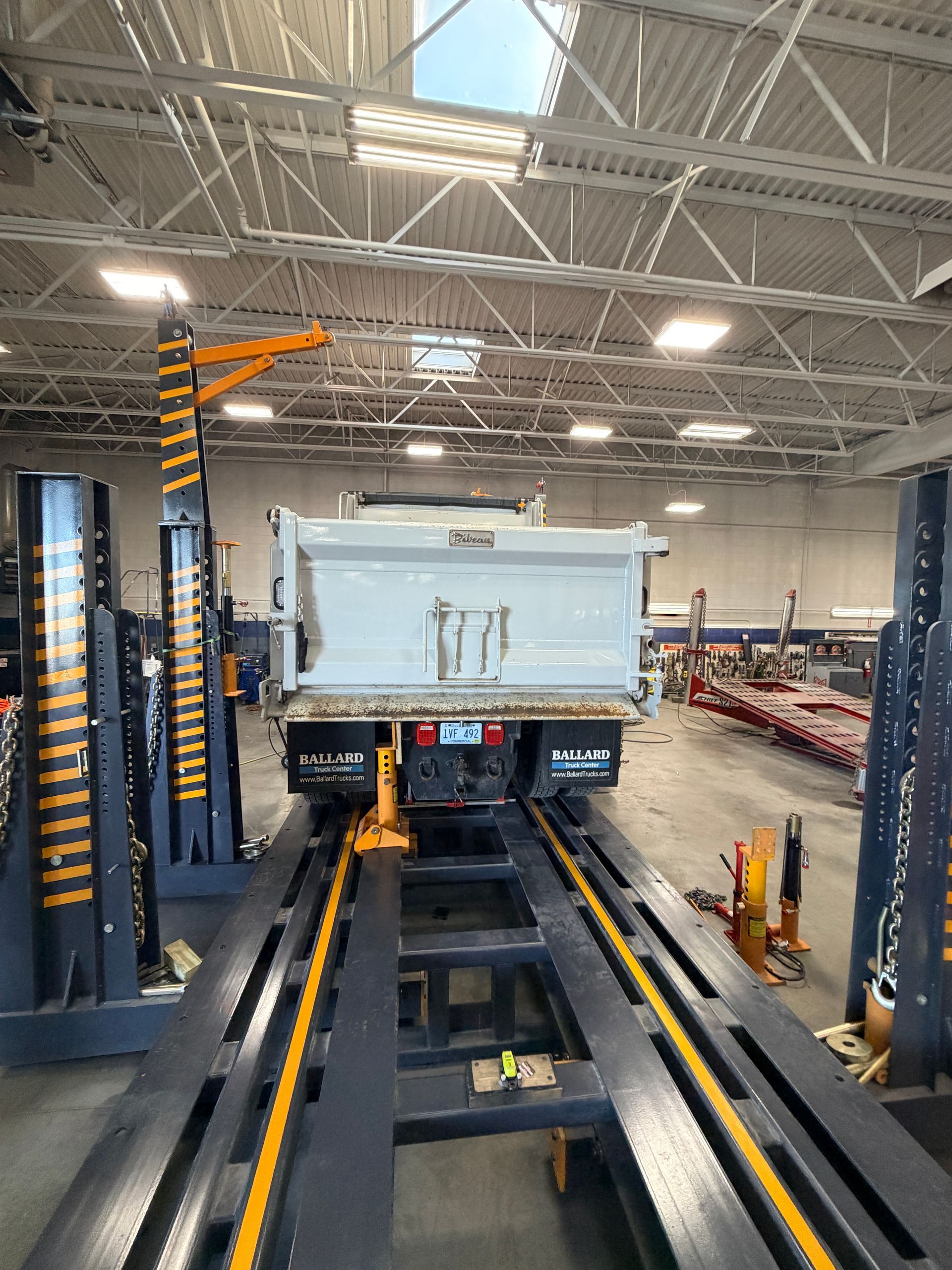 A white dump truck bed sits on a heavy-duty industrial lift system inside a maintenance garage.