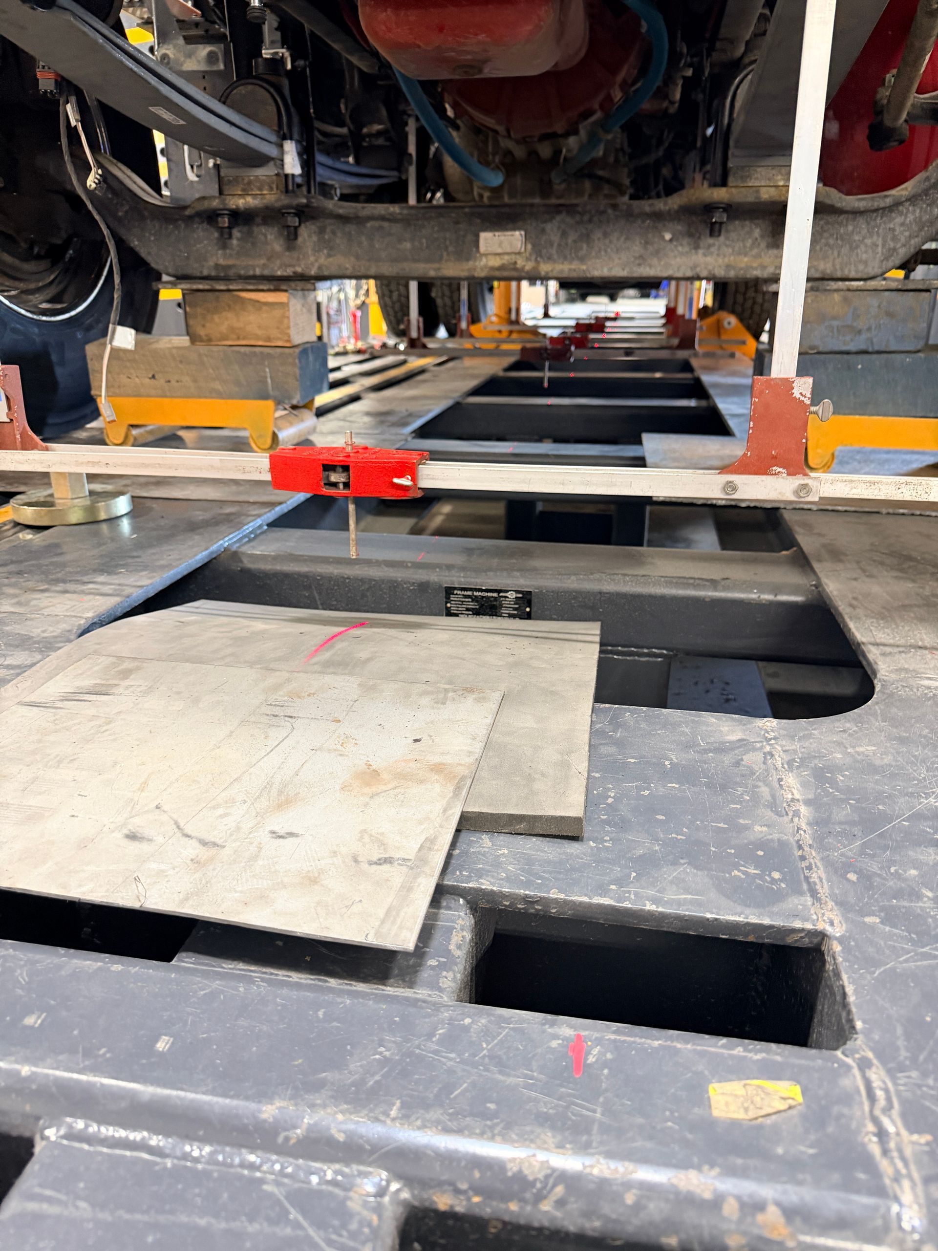 A red leveling tool sits on a bar across a metal frame under a vehicle chassis in a maintenance shop.
