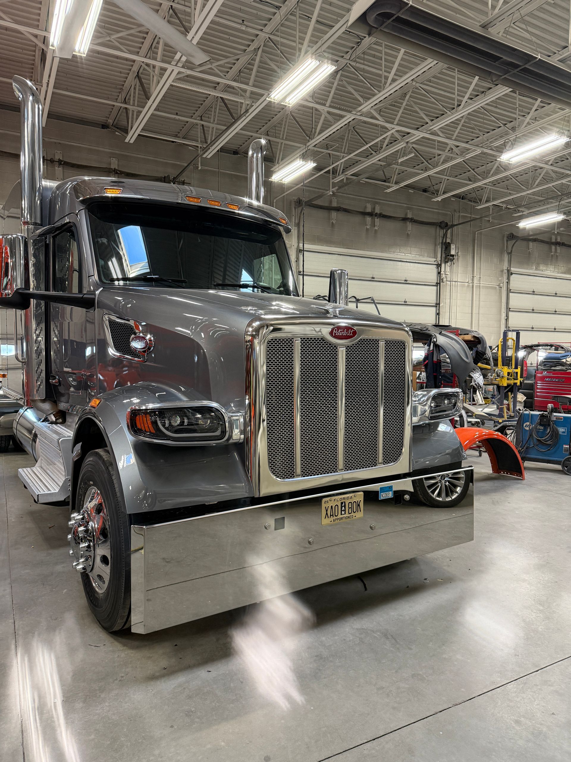 A grey semi-truck with a large chrome grille and bumper parked inside a maintenance workshop.