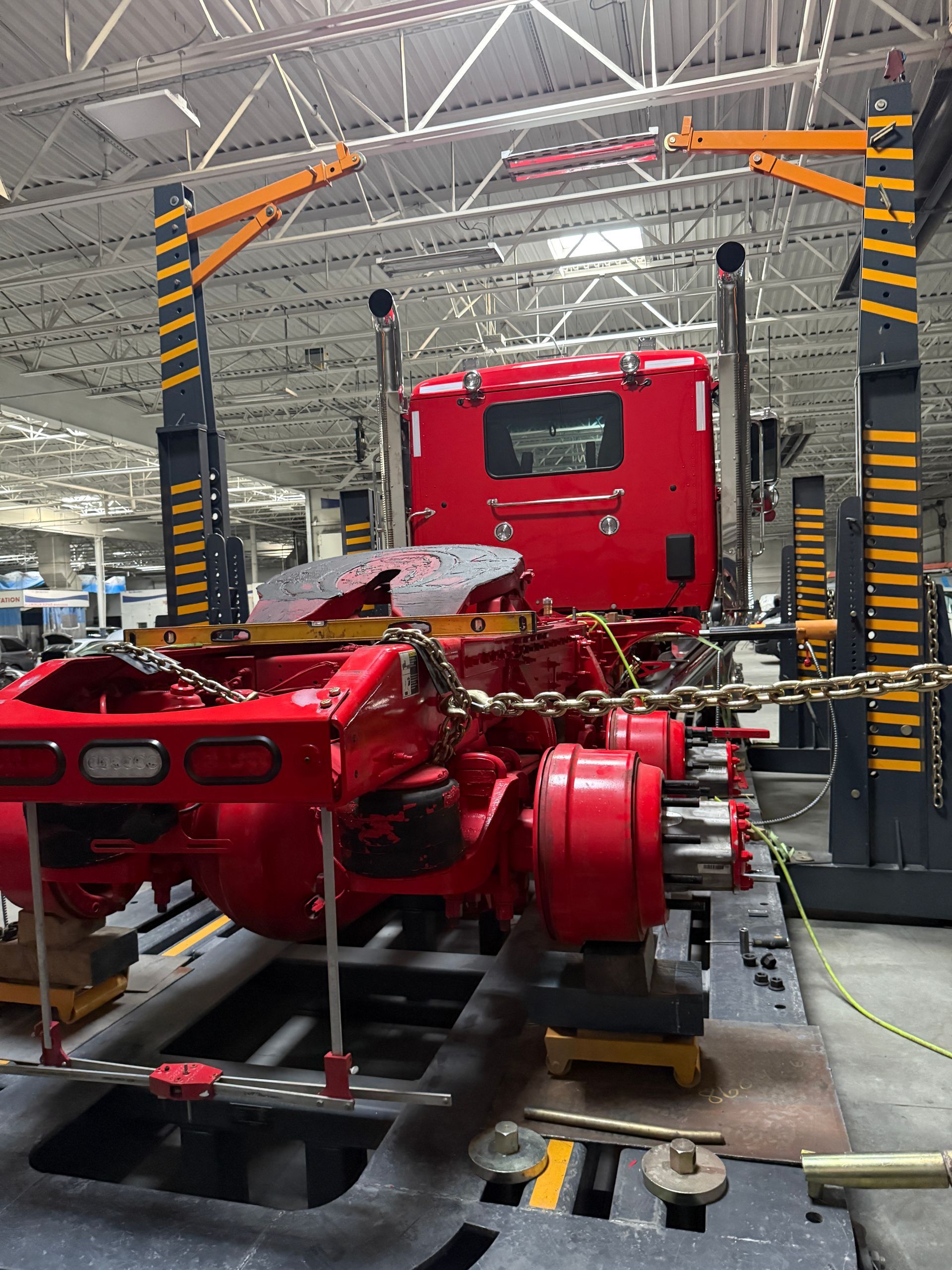 A bright red semi-truck frame is elevated on industrial heavy-duty lifts inside a brightly lit workshop.