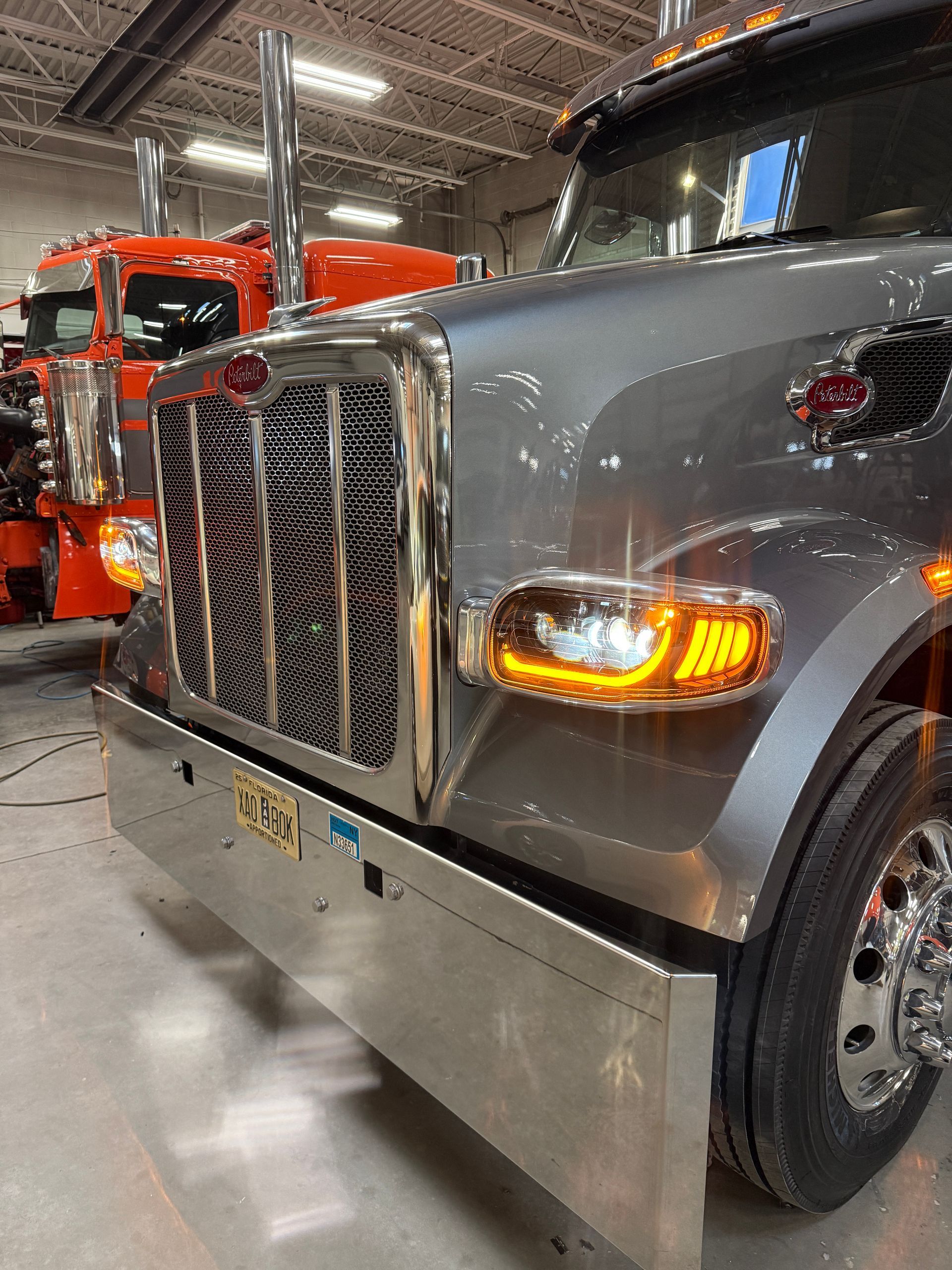 A close-up of a silver Peterbilt semi-truck parked indoors, with a portion of an orange truck visible in the background.