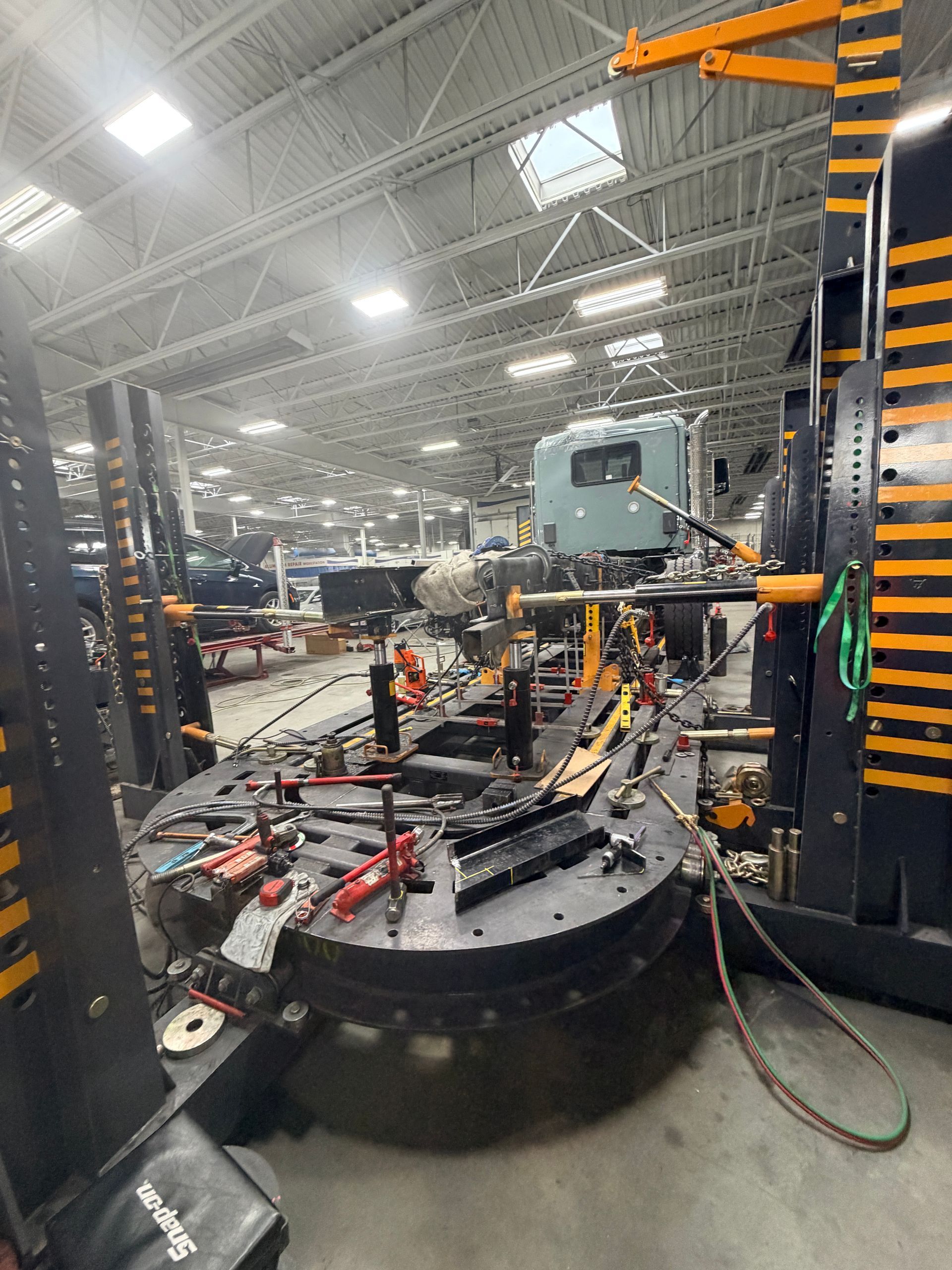 An automotive frame repair rack with a truck cab in a large, industrial workshop with bright overhead lighting.