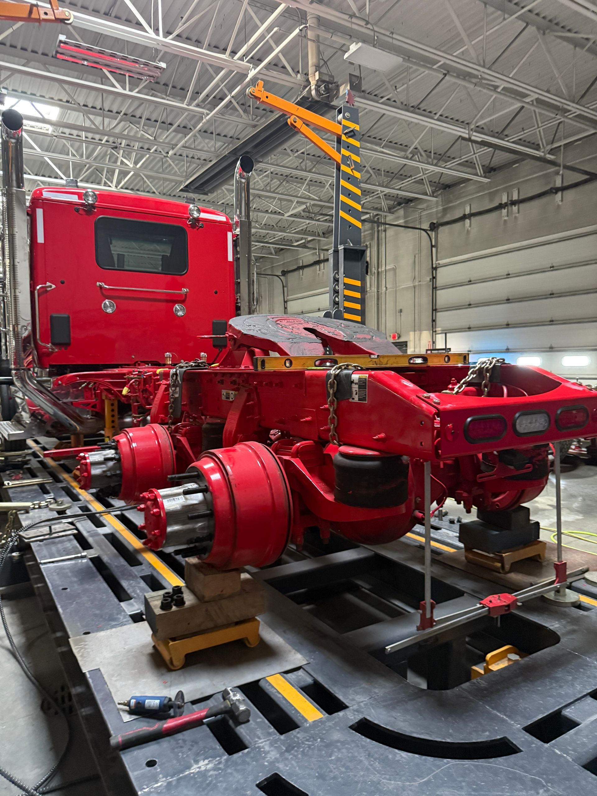 A red semi-truck cab frame being serviced on a raised platform in a workshop, with exposed axles and wheels.
