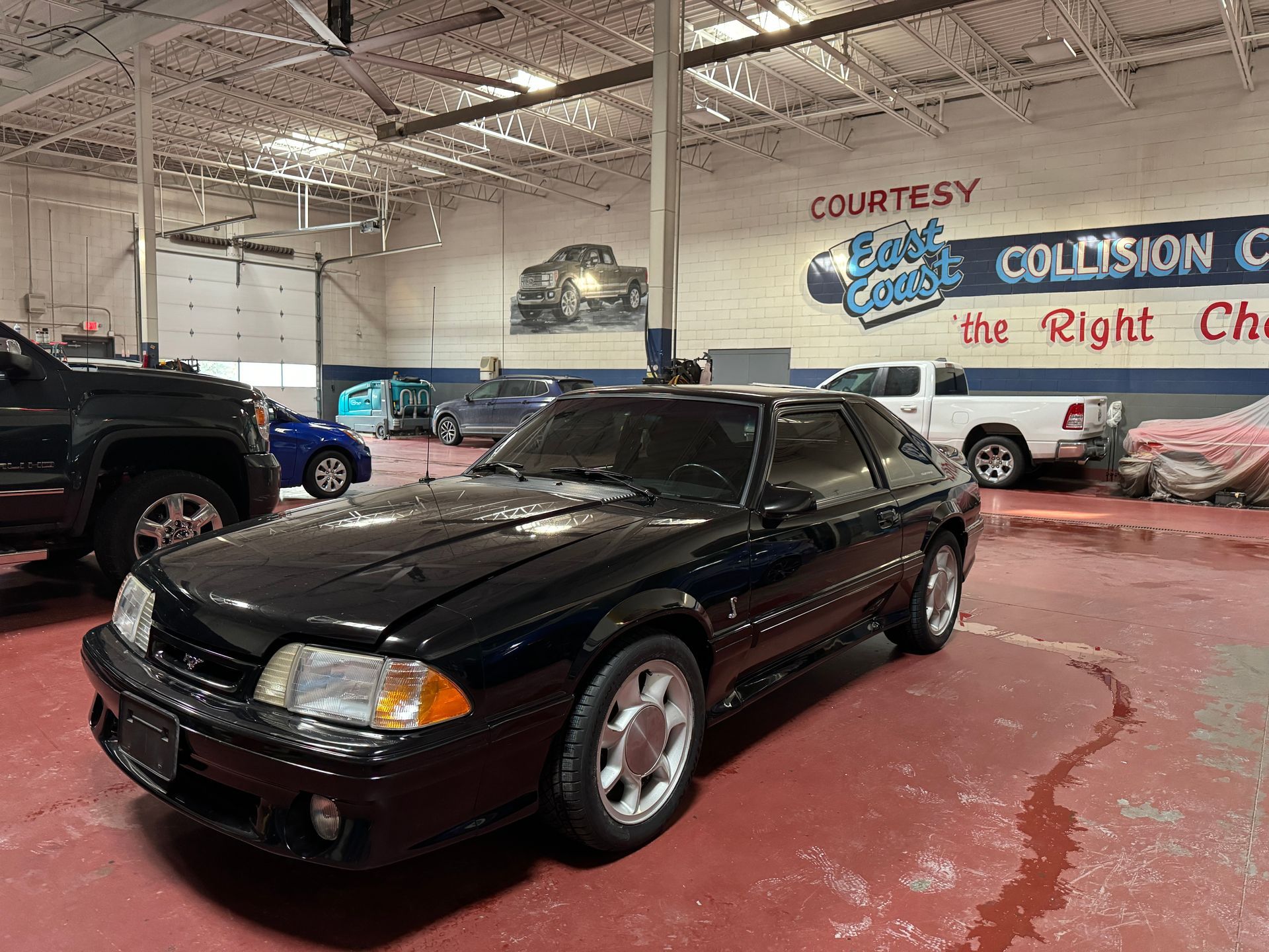A black Ford Mustang Fox Body parked in a vehicle repair shop with a white truck and other cars in the background.