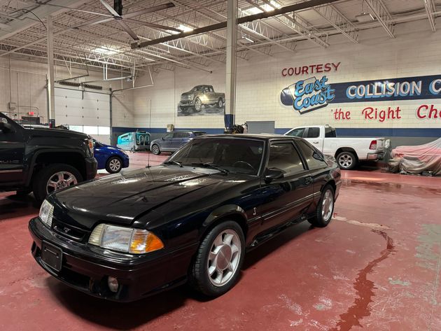 A black Ford Mustang Fox Body parked inside a spacious auto collision center with a large mural on the back wall.