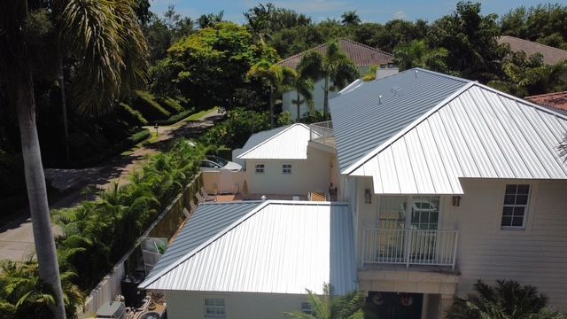 An aerial view of a white house with a metal roof surrounded by palm trees.