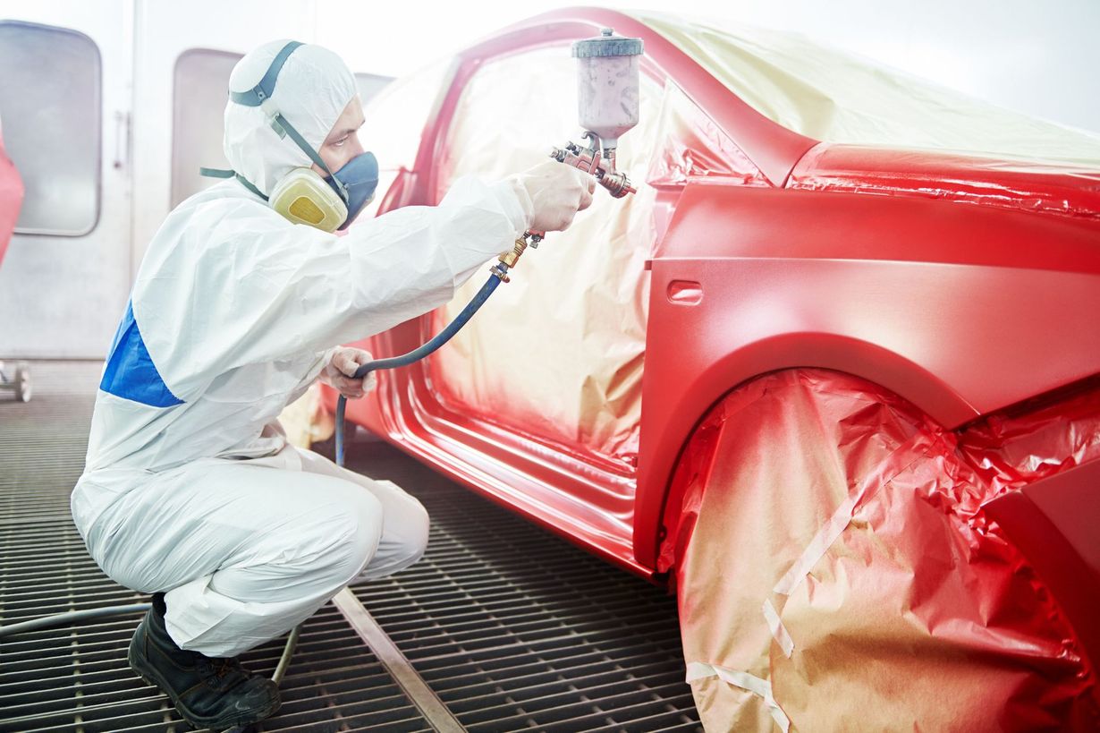 Person in protective suit painting a red car in a spray booth, using a spray gun.