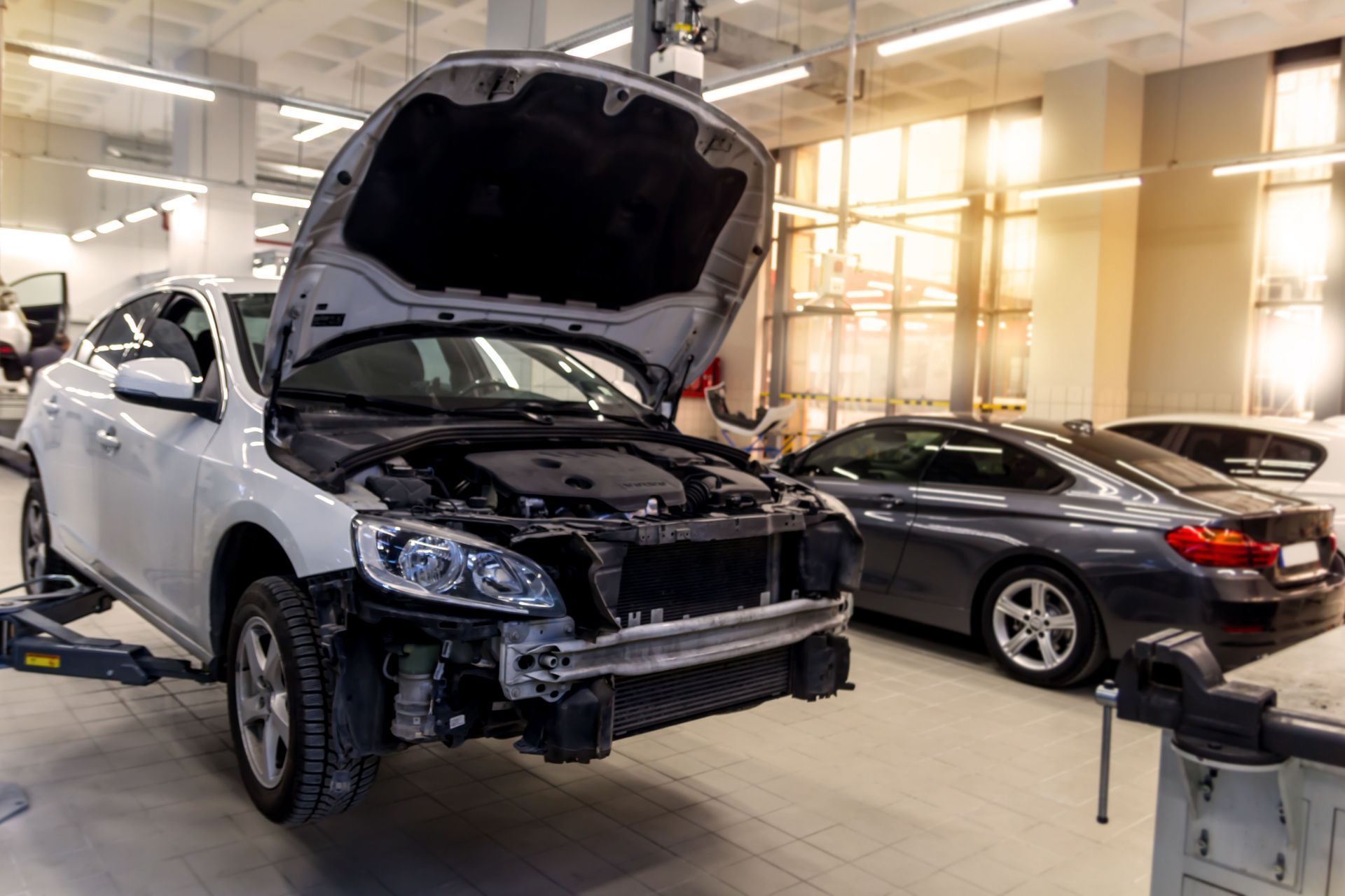 White car in a repair shop with hood open, front bumper removed. Another car in background.