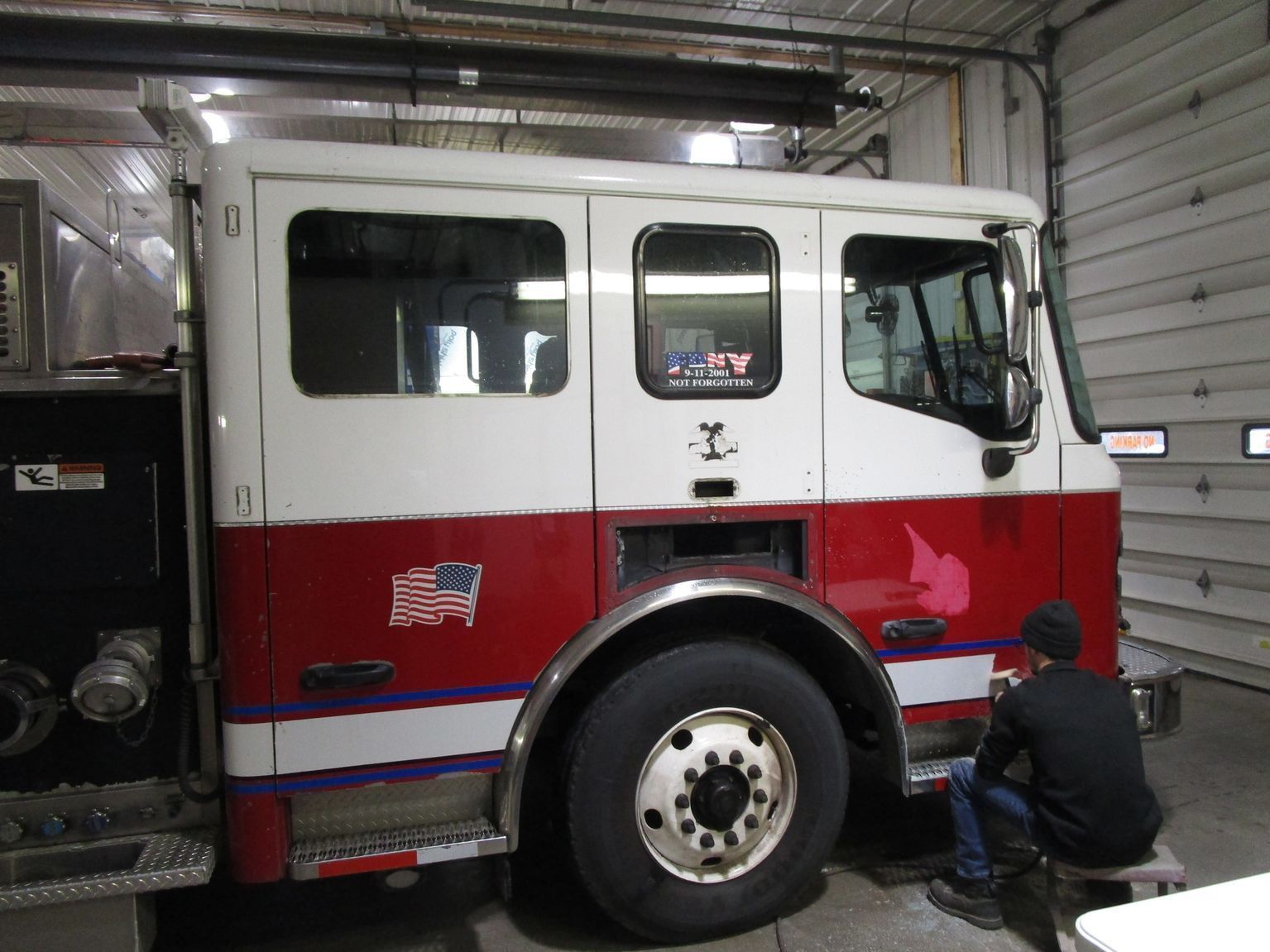 Fire truck, red and white, inside a garage with a person sitting on a stool near the wheel.