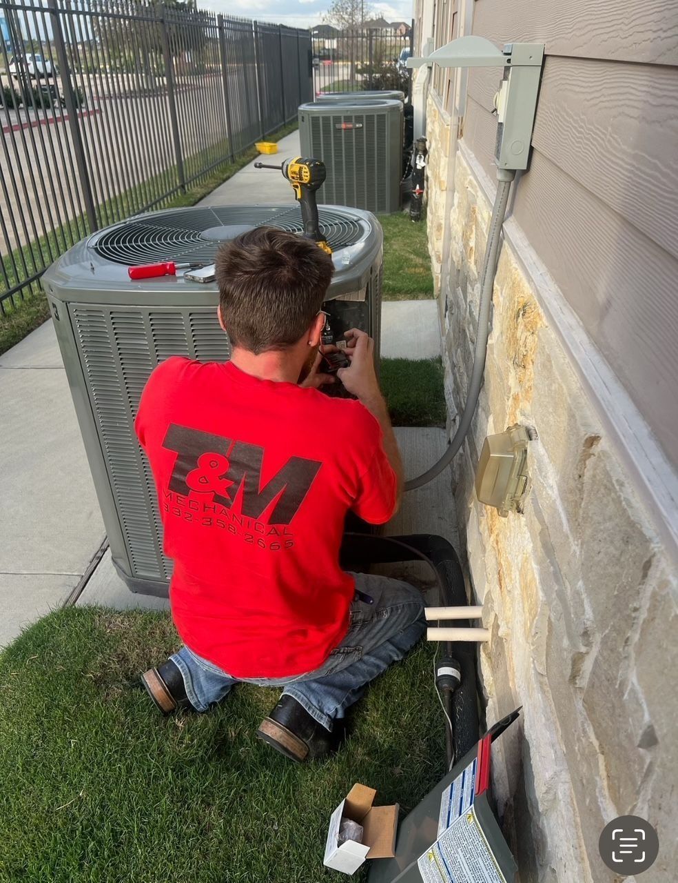 HVAC technician in red shirt repairing an air conditioning unit outside a building with stone siding.