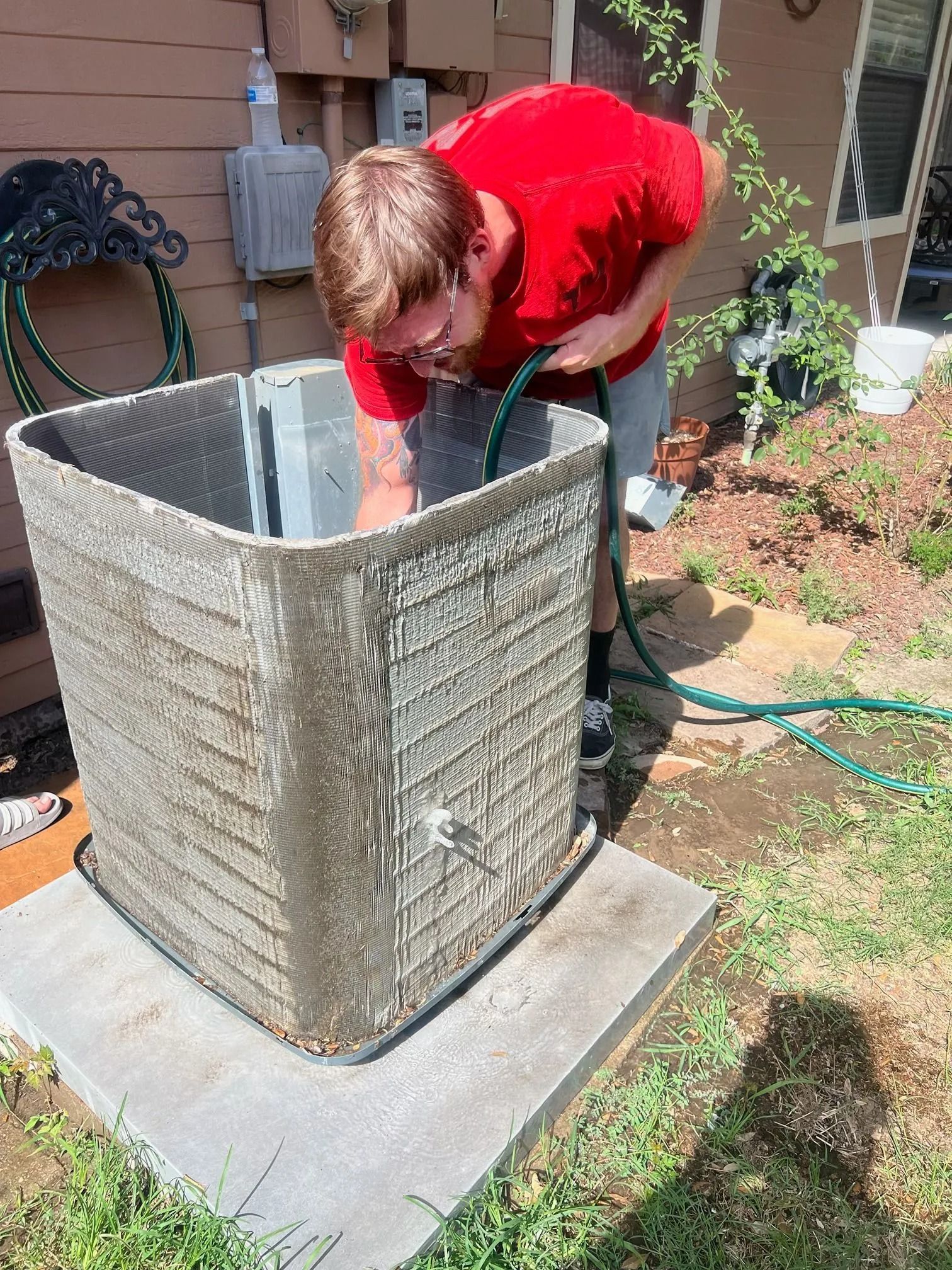 Man in red shirt sprays outdoor air conditioning unit with a hose.
