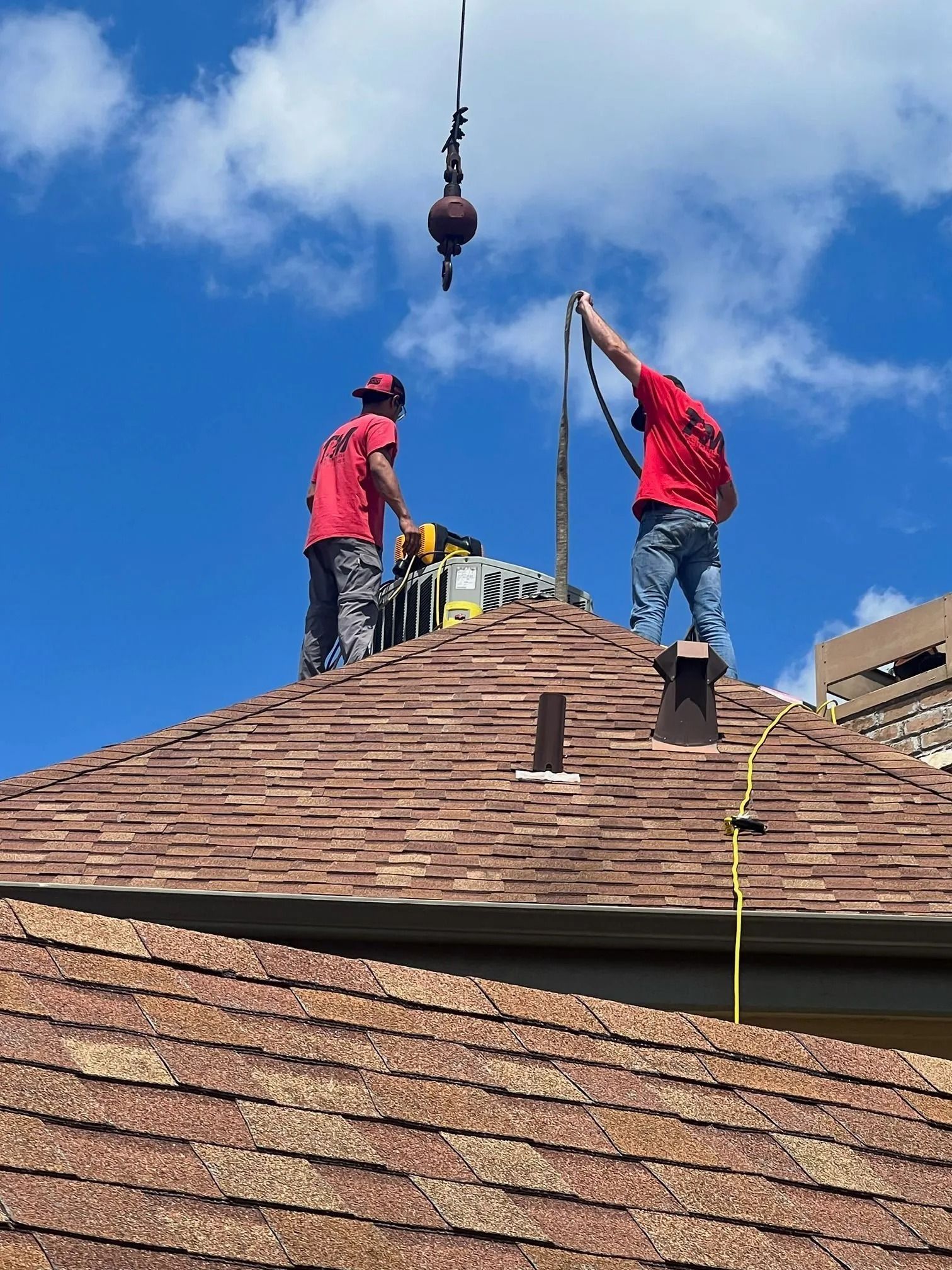 Two workers on a brown shingle roof, one holding a cable, another operating a saw, crane overhead, blue sky.