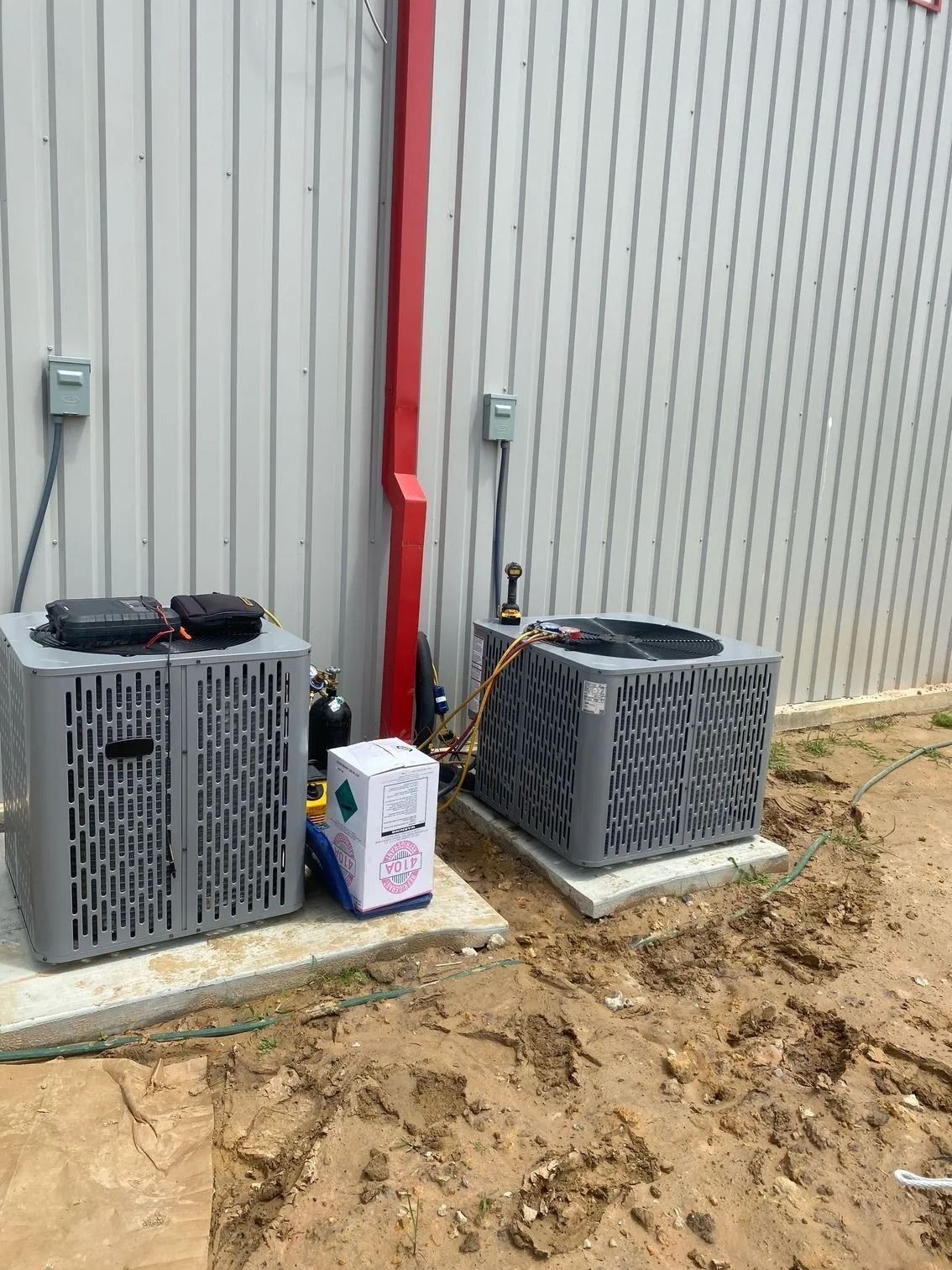 Two gray air conditioning units next to a white box and a red pipe against a white corrugated building.