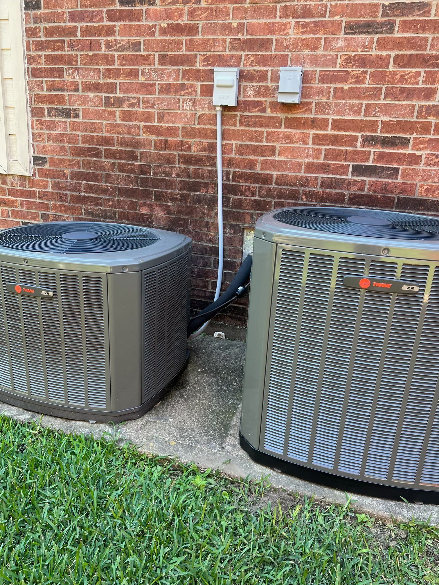 Two Trane air conditioning units sit on concrete next to a brick wall.