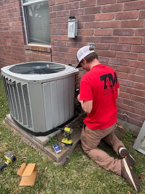 A person in red shirt kneels next to an AC unit, working on it near a brick wall.
