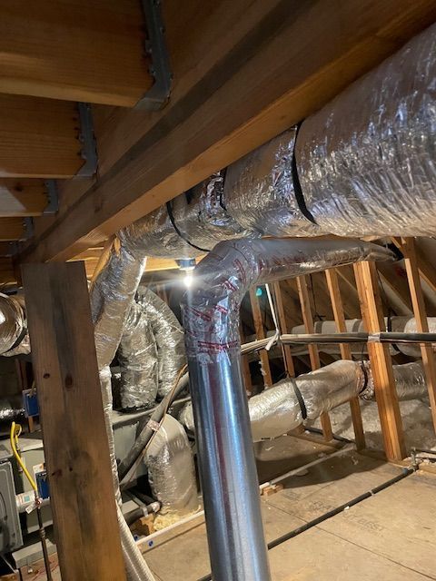 HVAC ductwork in an attic, with insulated flex duct connected to a metal vent pipe, amid wooden framing.