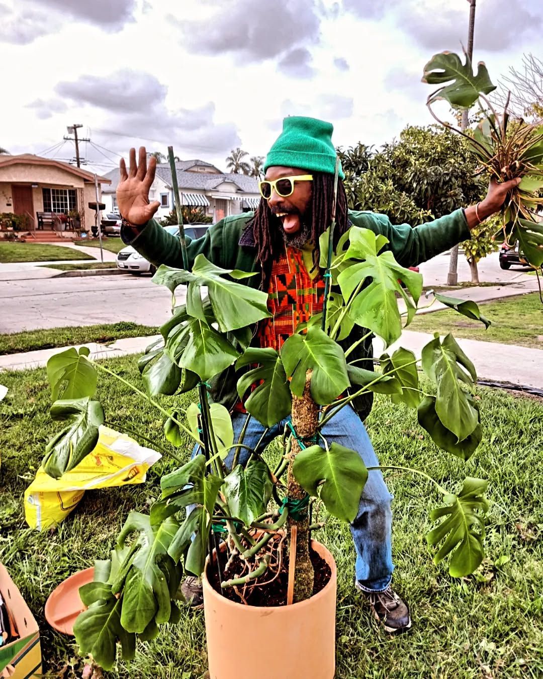 Man in green hat cheers by a potted Monstera plant outdoors.