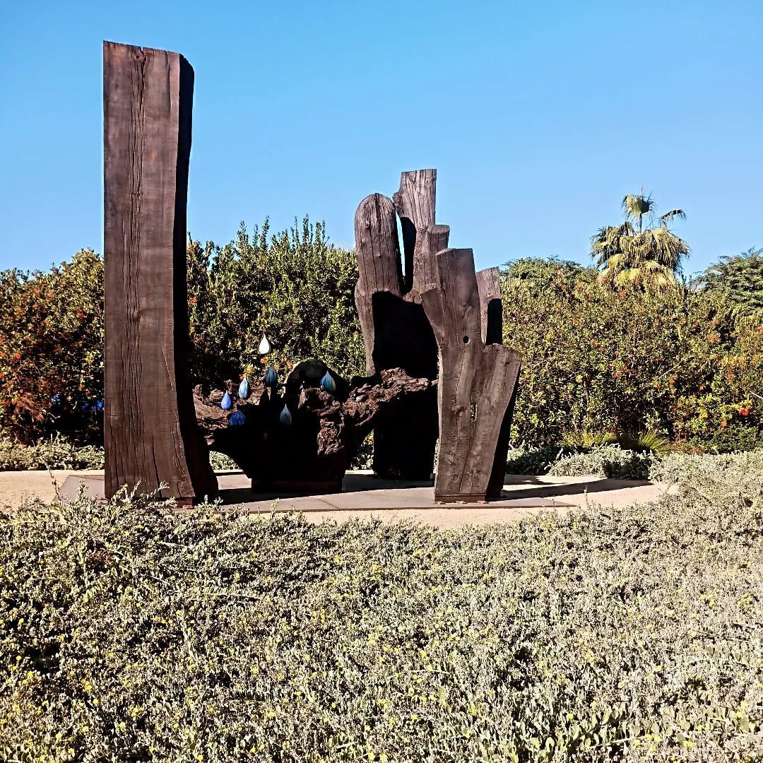 Wooden abstract sculpture in a garden setting, tall, dark brown forms against greenery and a blue sky.