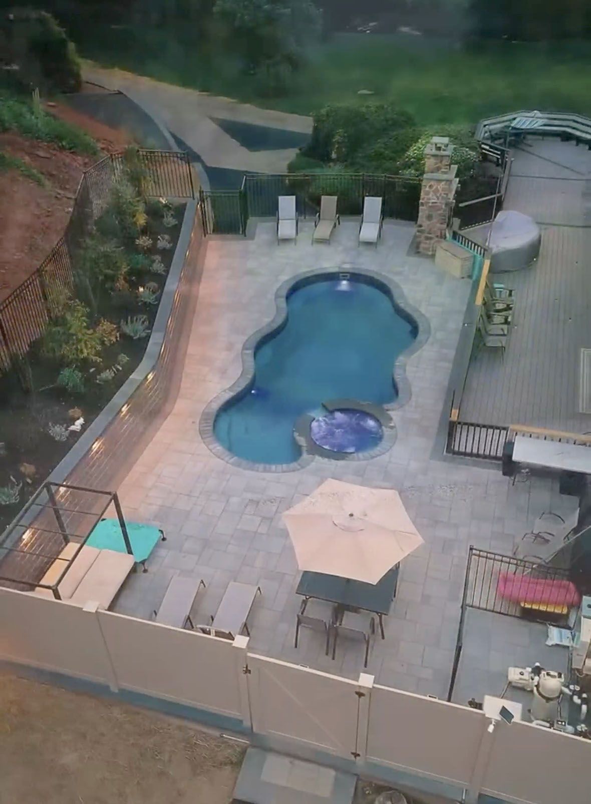Backyard swimming pool with patio chairs, umbrellas, and a hot tub beside a landscaped wall