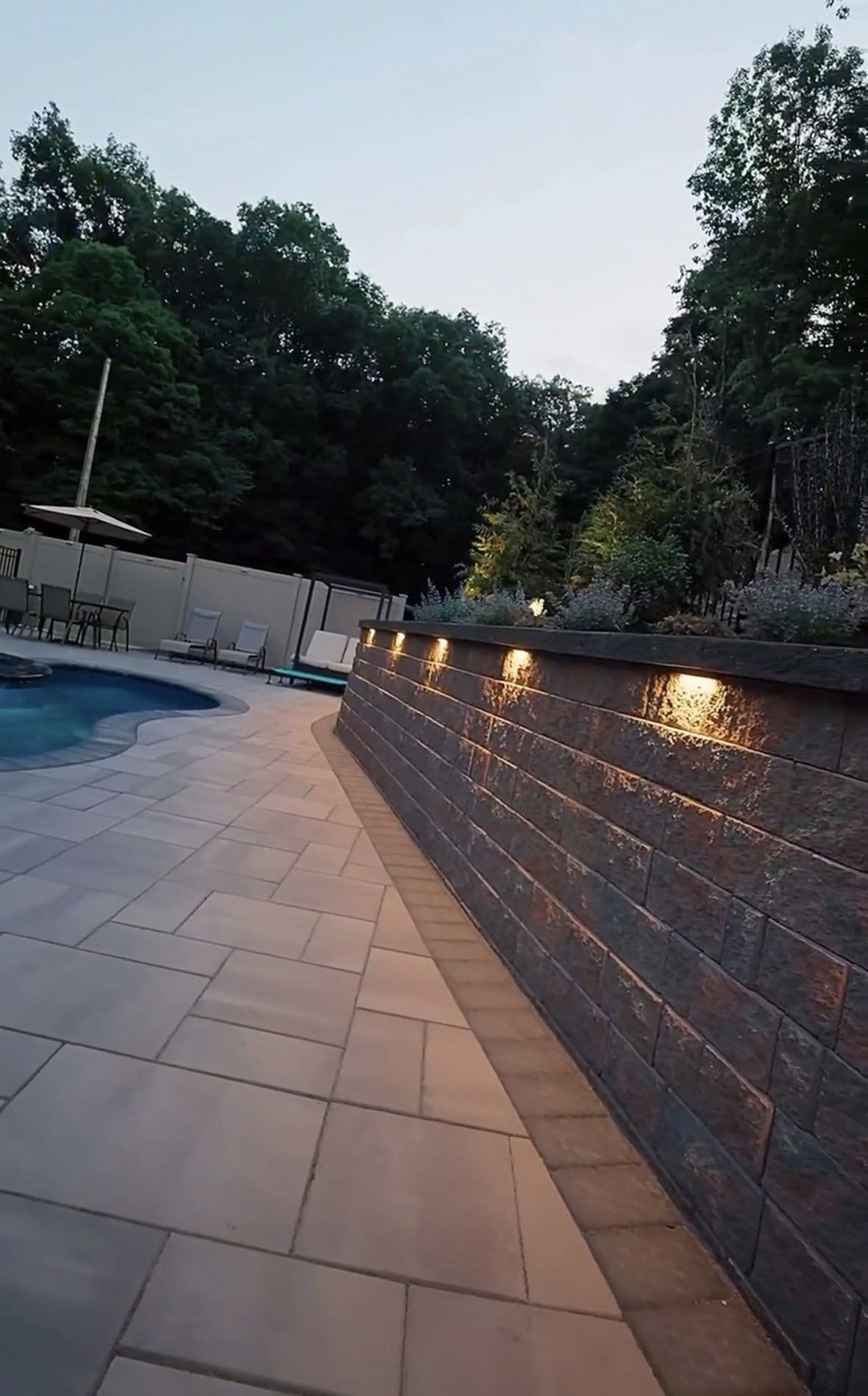 Lit poolside walkway beside a low stone wall with warm lights and trees at dusk
