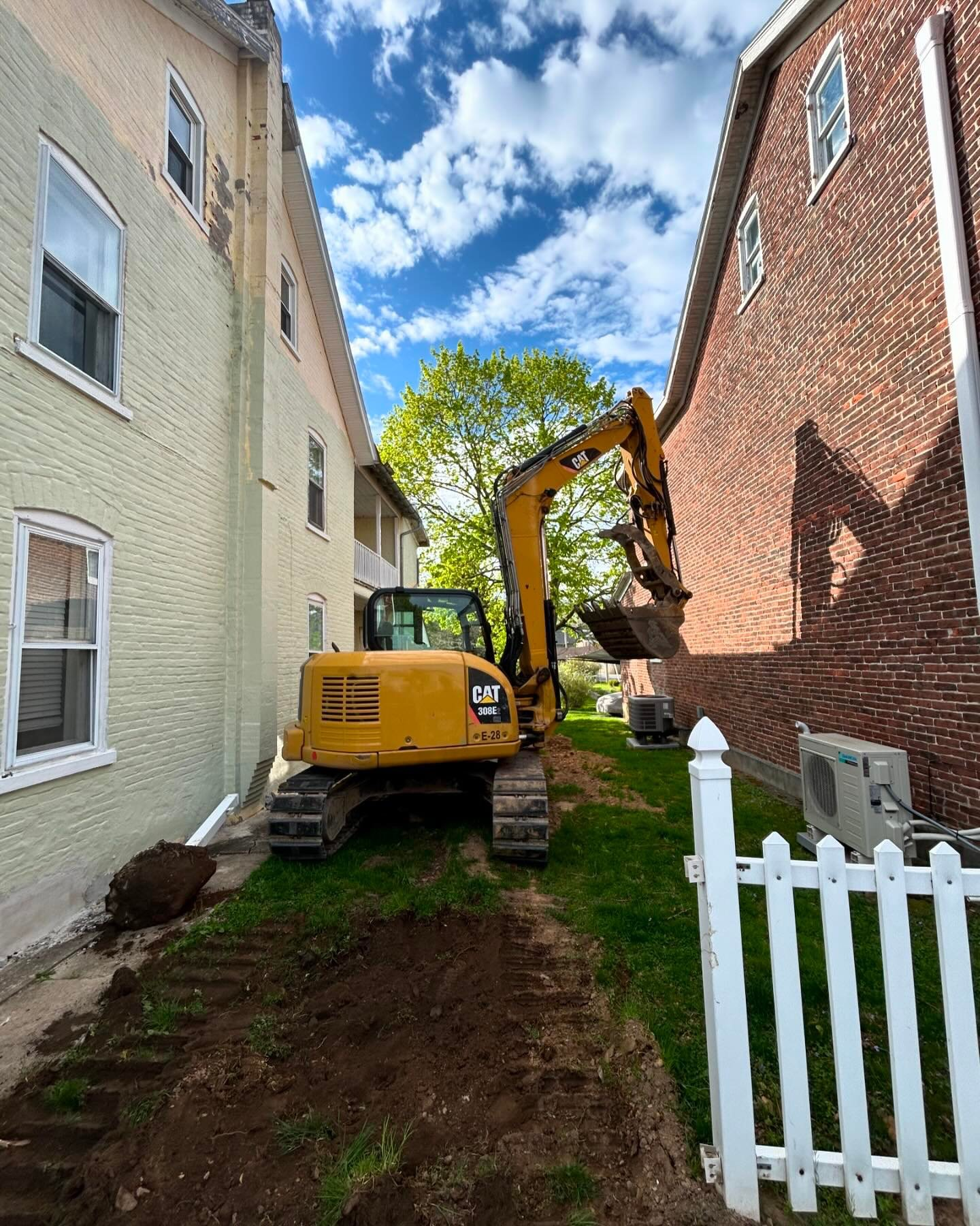 A yellow excavator is digging a hole in a yard between two buildings.