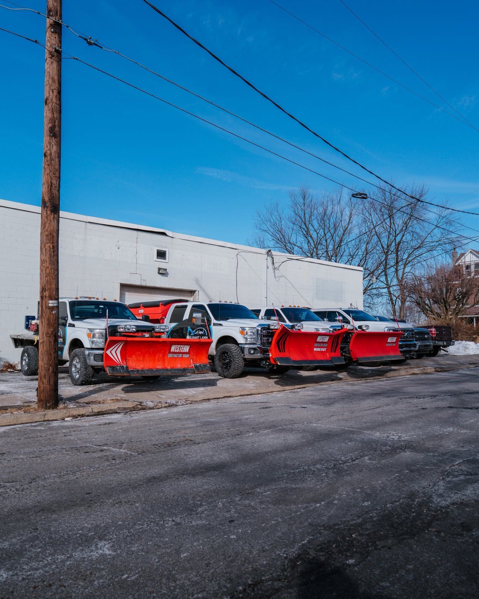 Snowplow trucks lined up in front of a white building on a sunny day.
