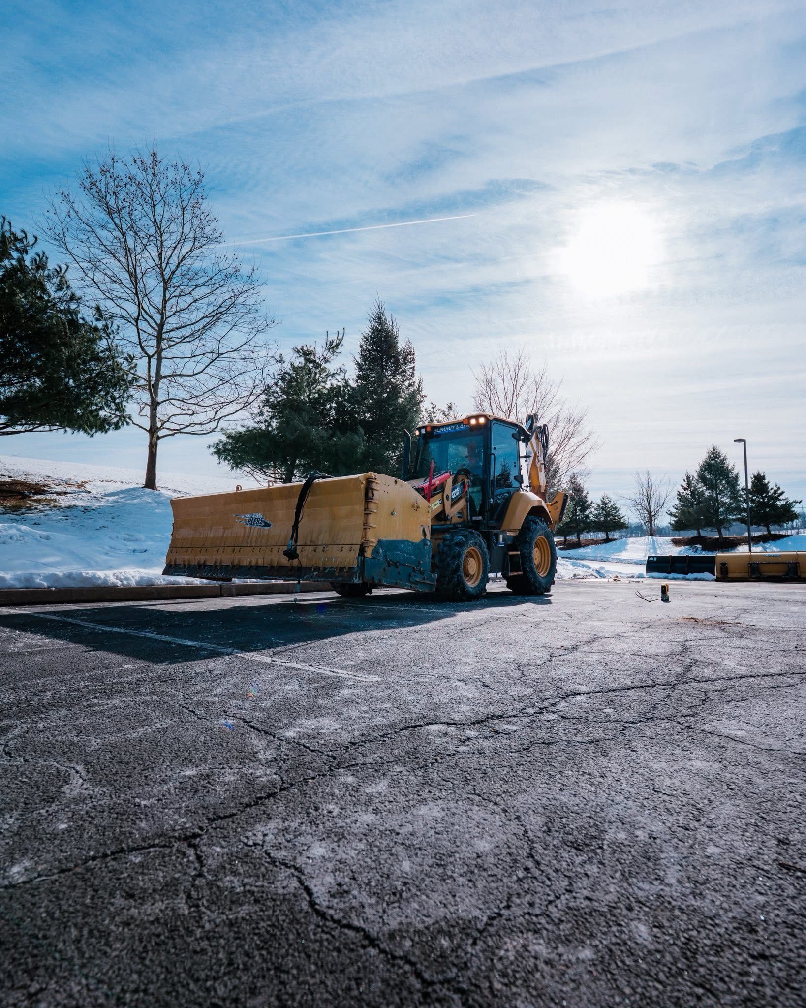 Yellow snowplow clearing snow on a parking lot under a bright sun.