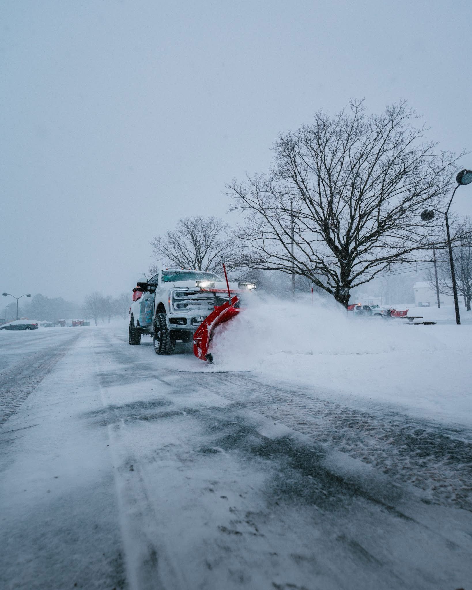 Snowplow clearing a snow-covered road during a winter storm, with trees and a parking area in the background.